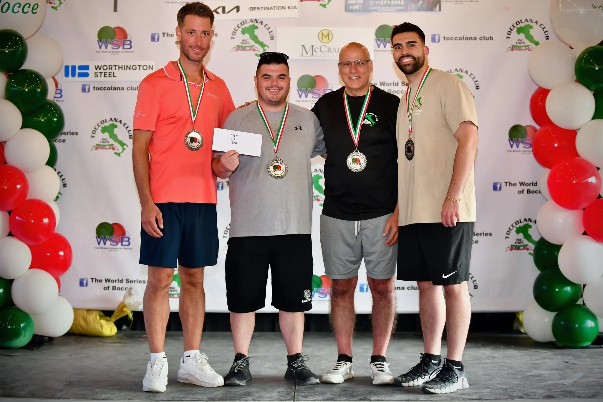 Four men standing on a stage with bocce medals around their necks, smiling for a photo. One man is holding a check. The background has balloons and banners promoting the Toccolana Club and World Series of Bocce.