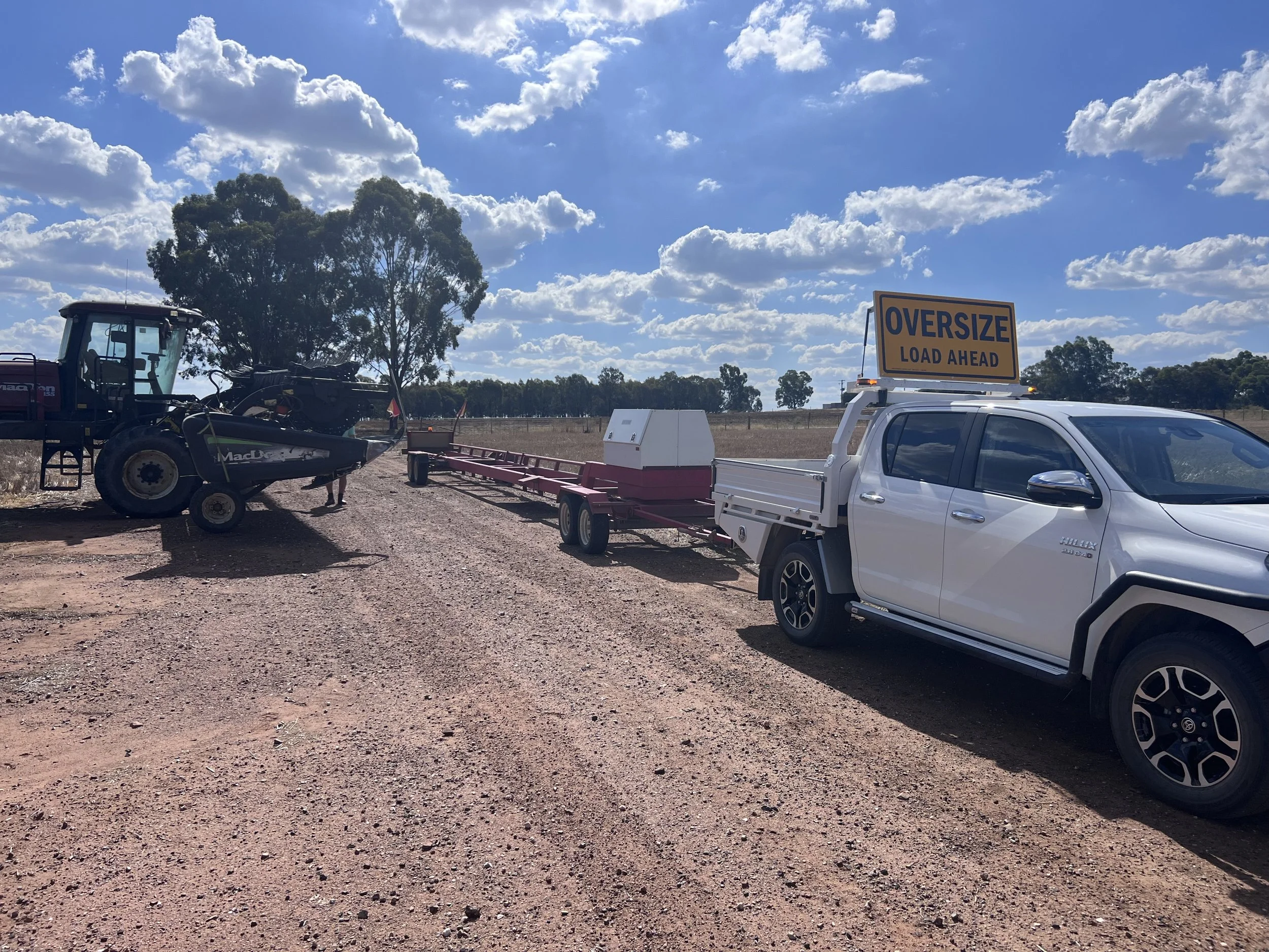 A white pickup truck with a yellow 'OVERSIZE LOAD AHEAD' sign on top pulling a float trailer, parked on a dirt field with trees and blue sky with clouds in the background.