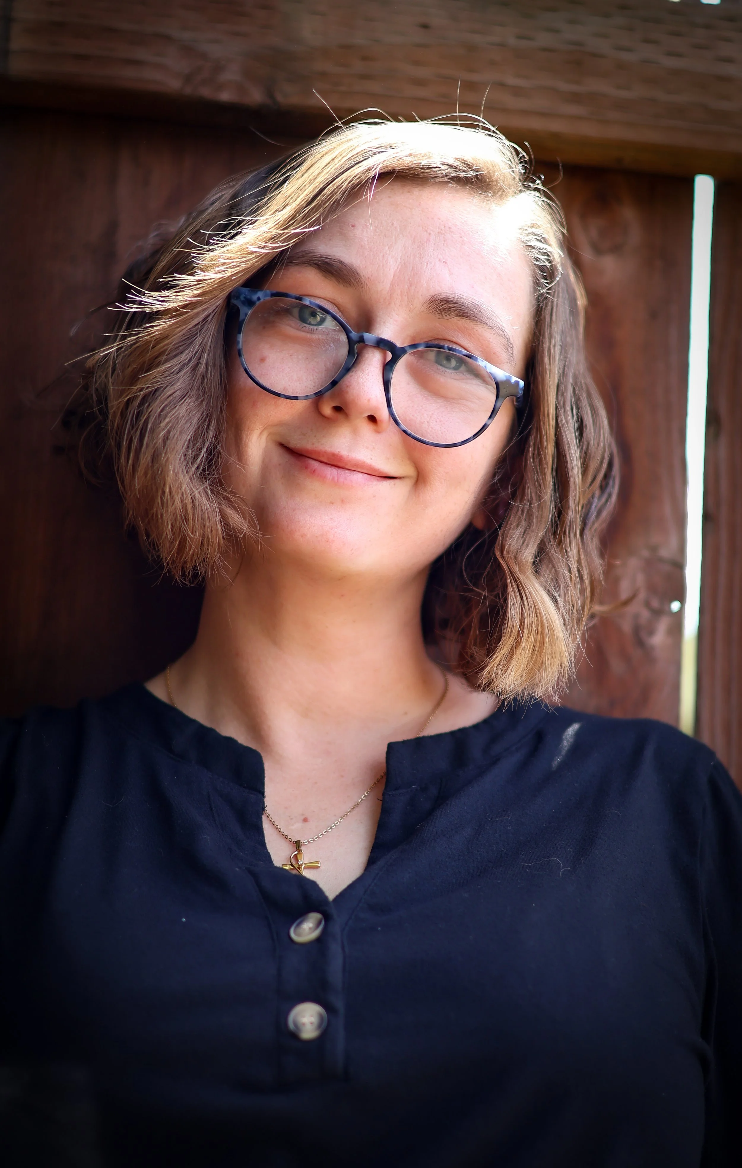 Close-up of a young woman with wavy, shoulder-length hair, wearing glasses with a tortoiseshell frame, smiling softly, wearing a dark shirt with buttons, and a gold necklace with a cross pendant, standing outdoors with a wooden background.