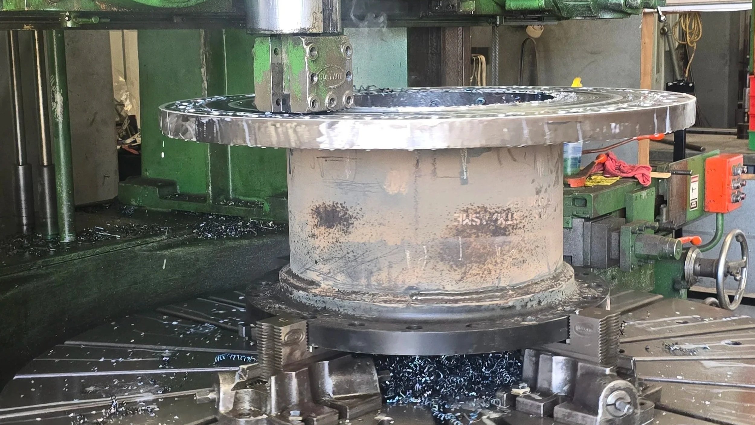 Close-up of a metal turning machine with a large, cylindrical workpiece held in place, surrounded by metal shavings and coolant liquid.