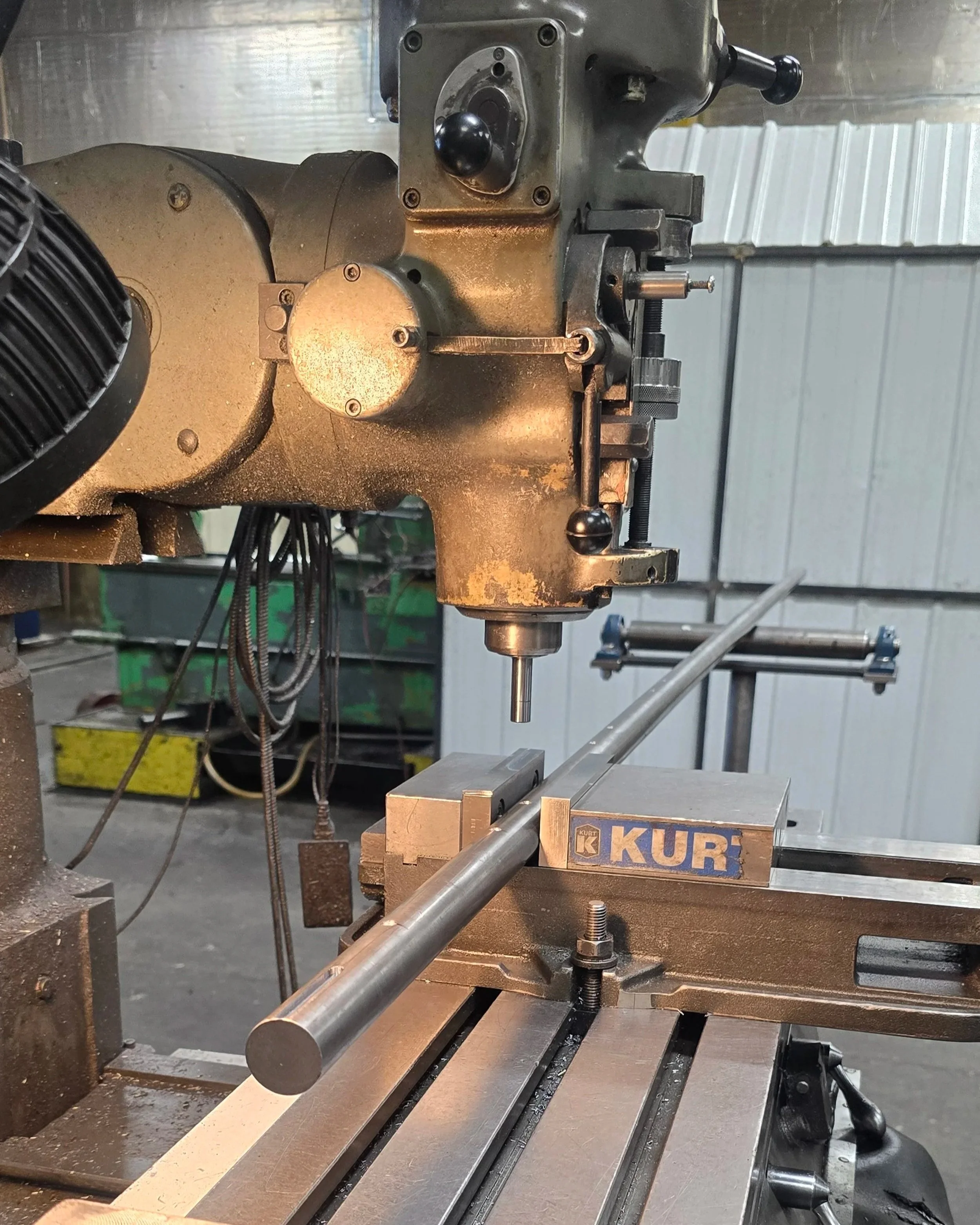 A metal lathe machine in a workshop, with a long metal rod being worked on, mounted on the lathe's bed, and the lathe's control handles and mechanisms visible.