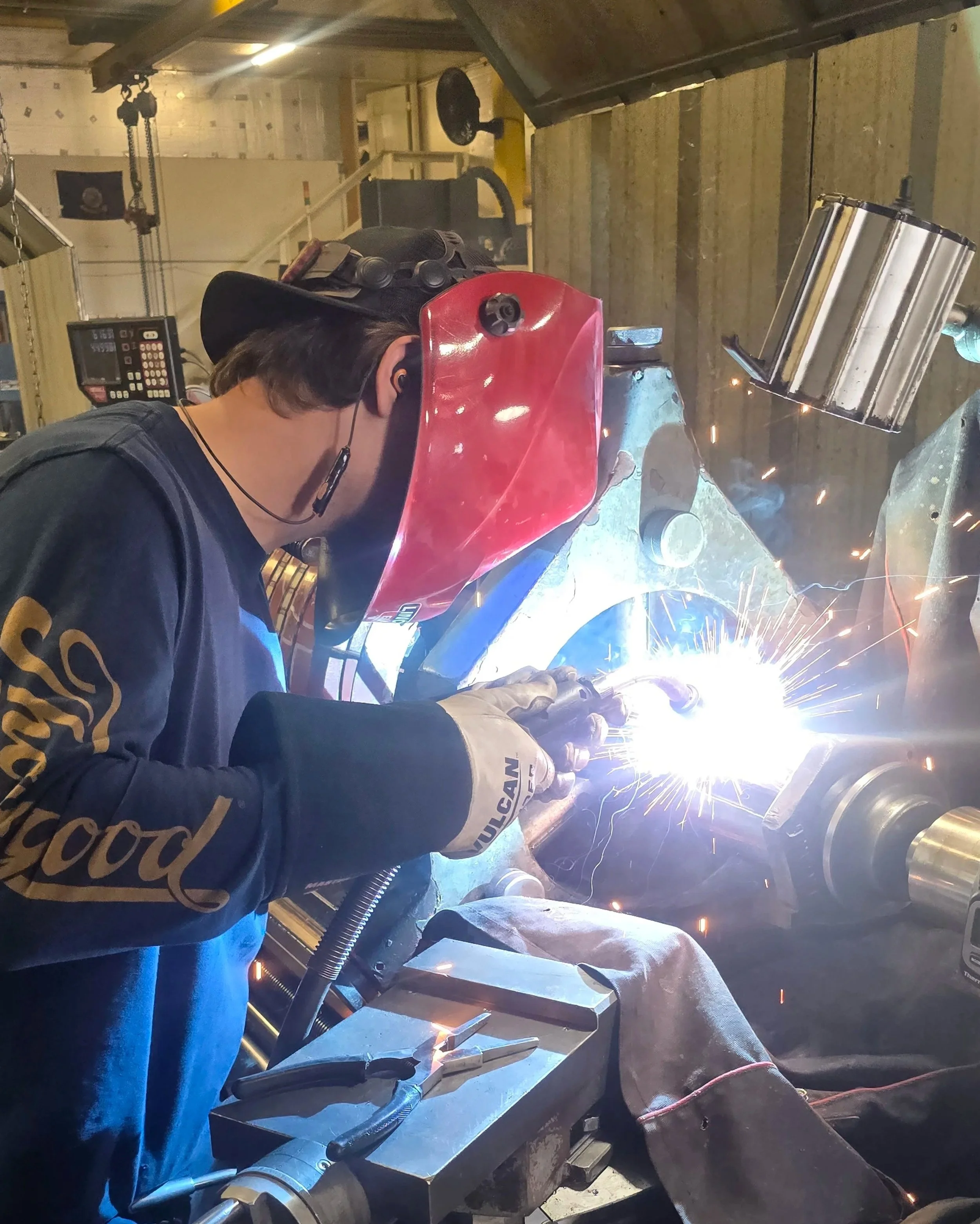 A person welding metal in a workshop, wearing a red welding helmet, gloves, and protective clothing, with sparks flying from the welding process.