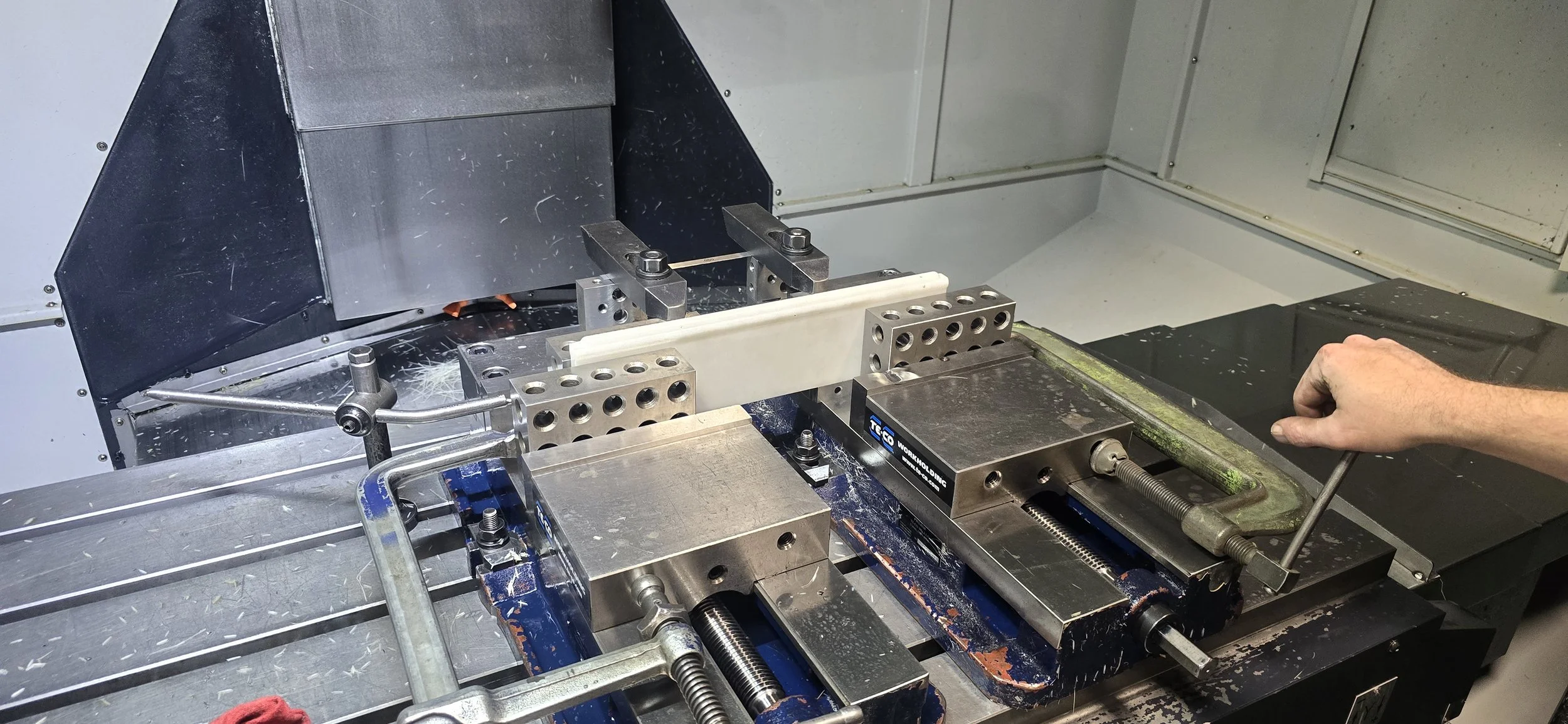 A CNC milling machine with metal clamps and a workpiece, operator's hand adjusting a screw.