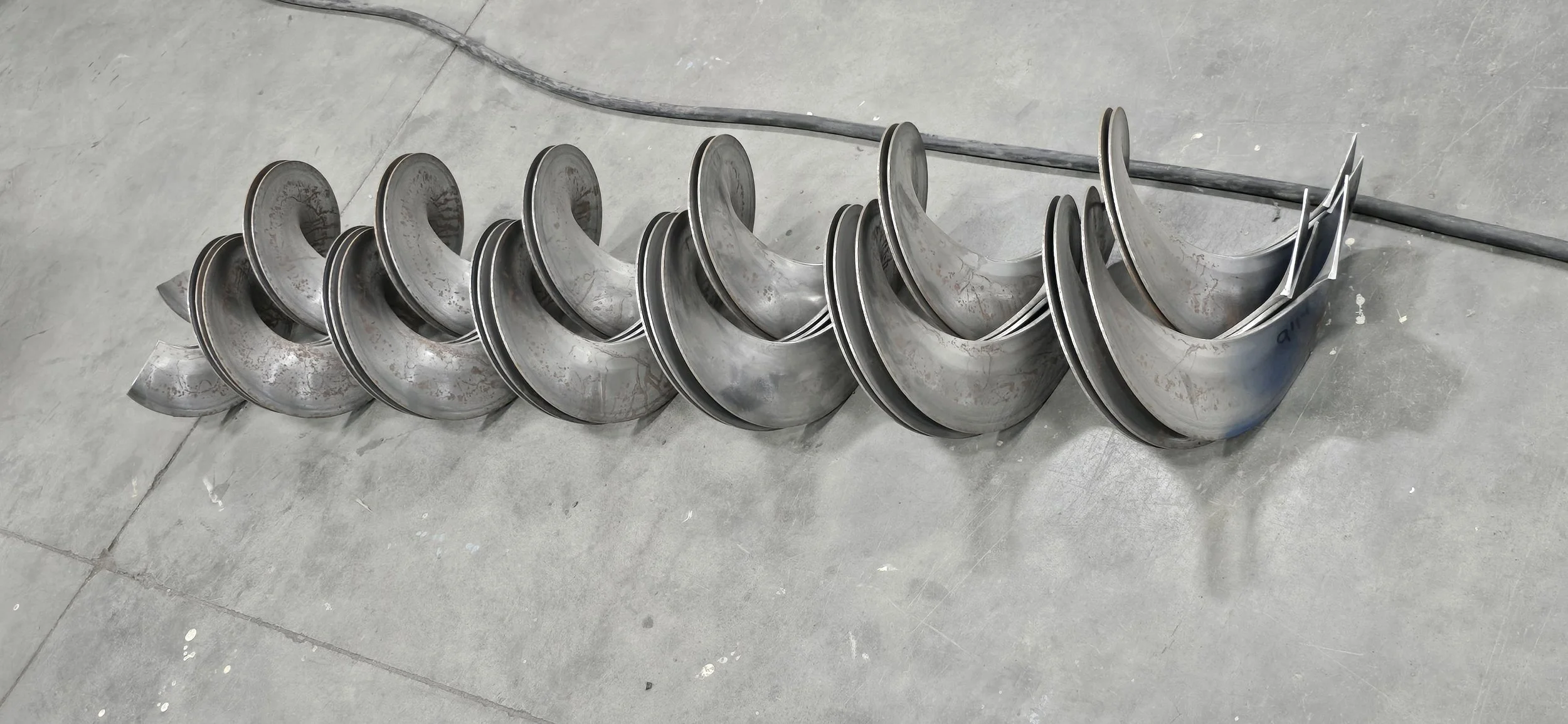 Large metal screw conveyor bent in a spiral shape and lying on a gray concrete floor.