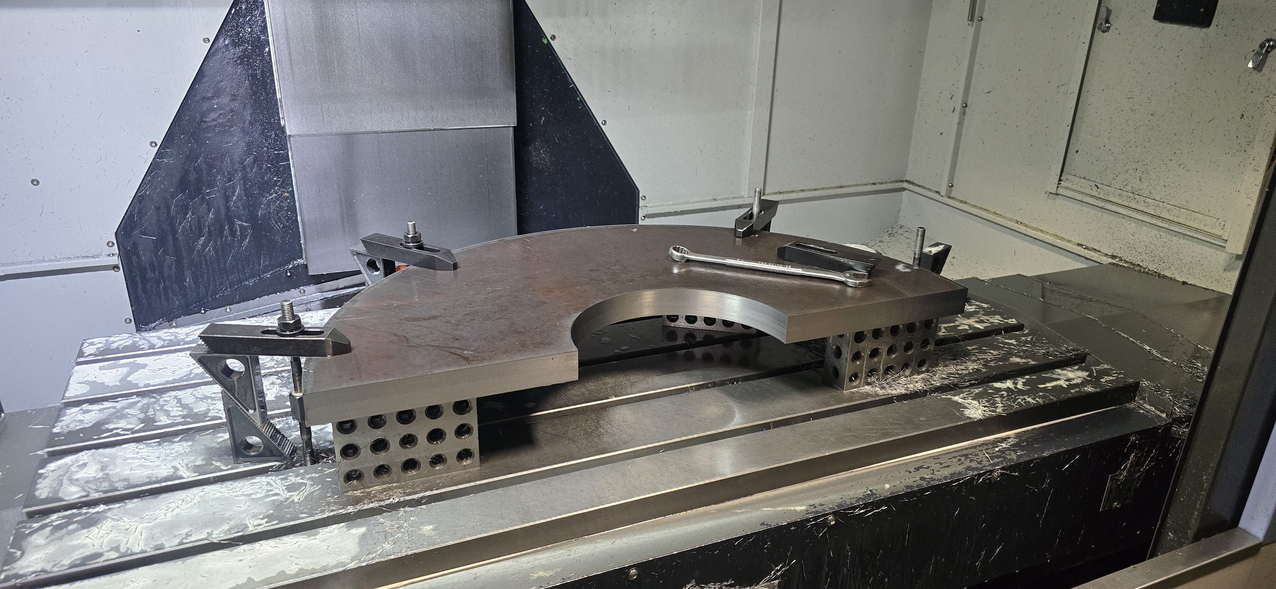 Metal workpiece secured on a machine bed with clamps in a machining center, with tools and fixtures visible around the workpiece.