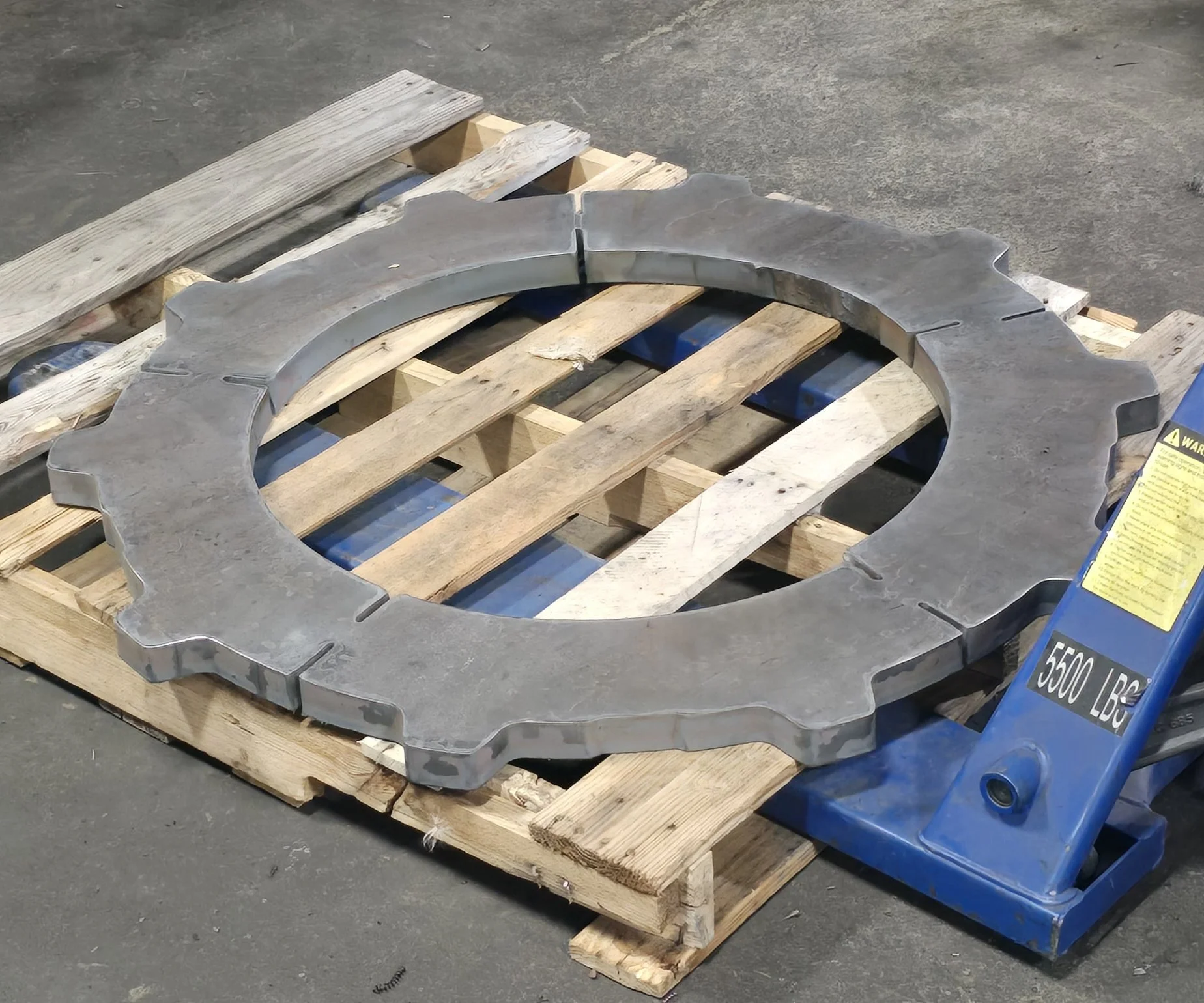 A large metal gear resting on a wooden pallet with a blue hydraulic lift nearby.
