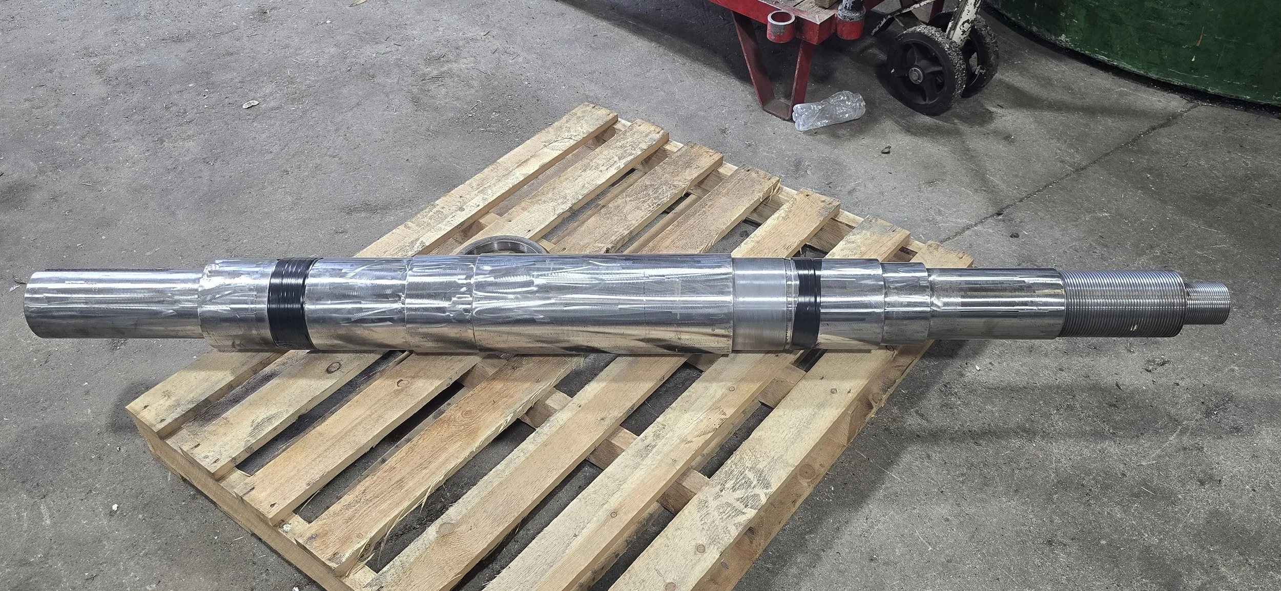 A large metal shaft with a polished surface, lying on a wooden pallet in an industrial setting.