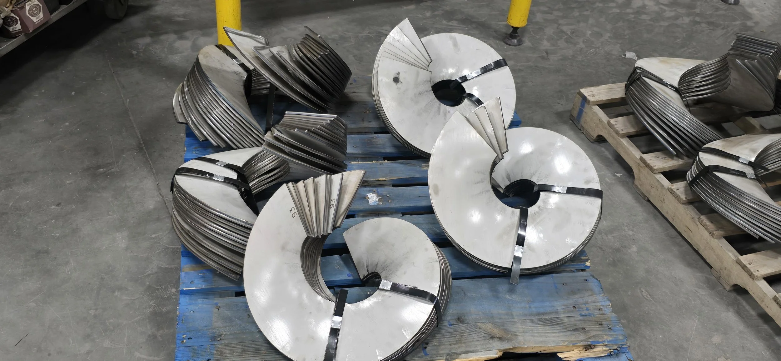 Metal components shaped in the form of large, circular, crescent-like structures, stacked and organized on a wooden pallet in an industrial setting.