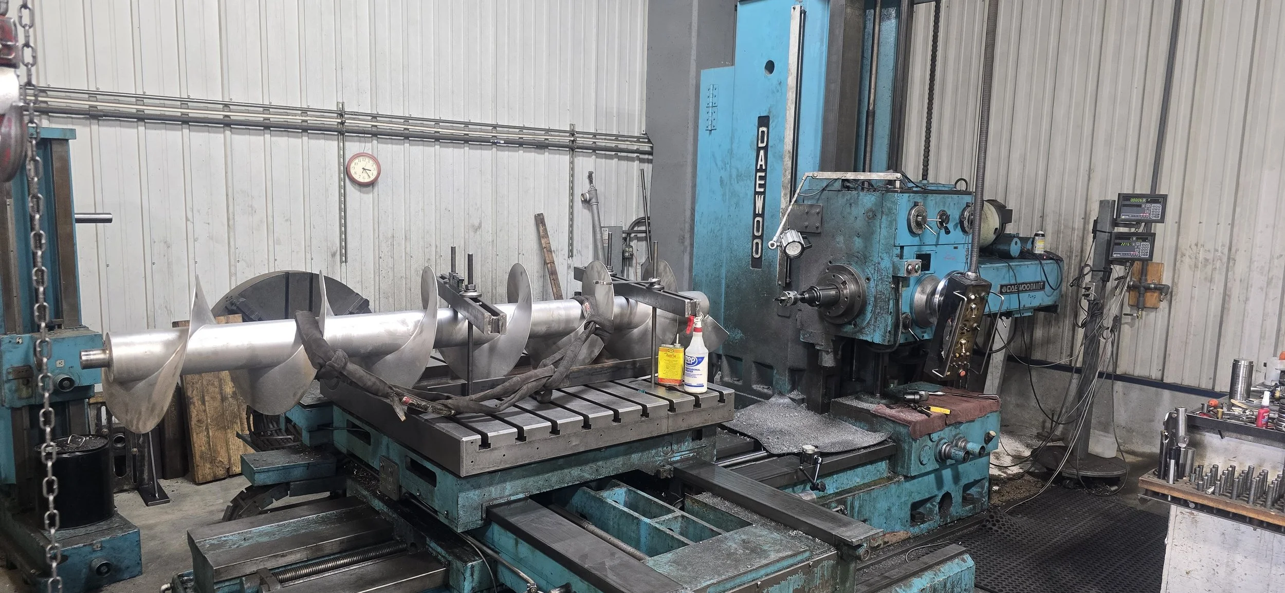Industrial workshop with a large blue machine, metal parts, tools, and cleaning supplies on a work table.