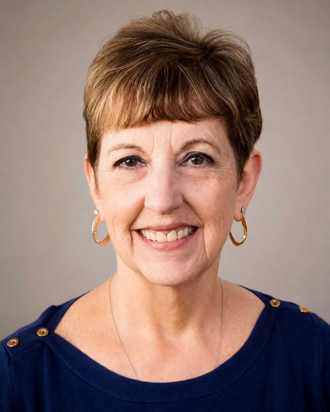 A smiling woman with short, brown hair, wearing gold hoop earrings and a navy blue top with gold button details on the shoulders.
