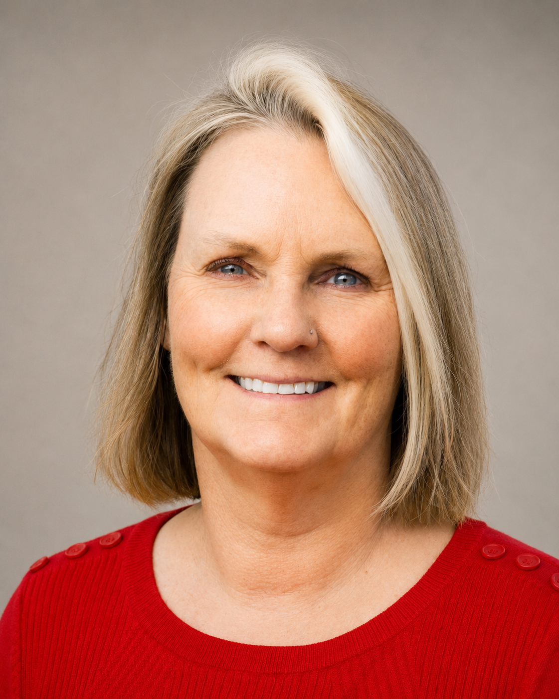 A woman with blonde hair and blue eyes smiling, wearing a red top with decorative buttons on the shoulders, against a neutral background.