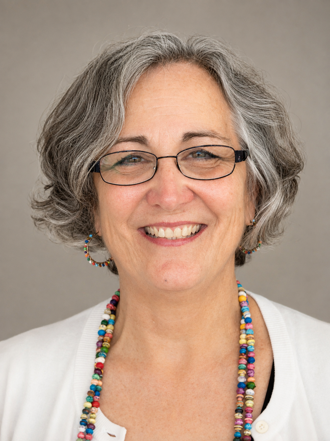 A smiling middle-aged woman with gray curly hair, wearing glasses, colorful beaded jewelry, a white shirt, and hoop earrings.