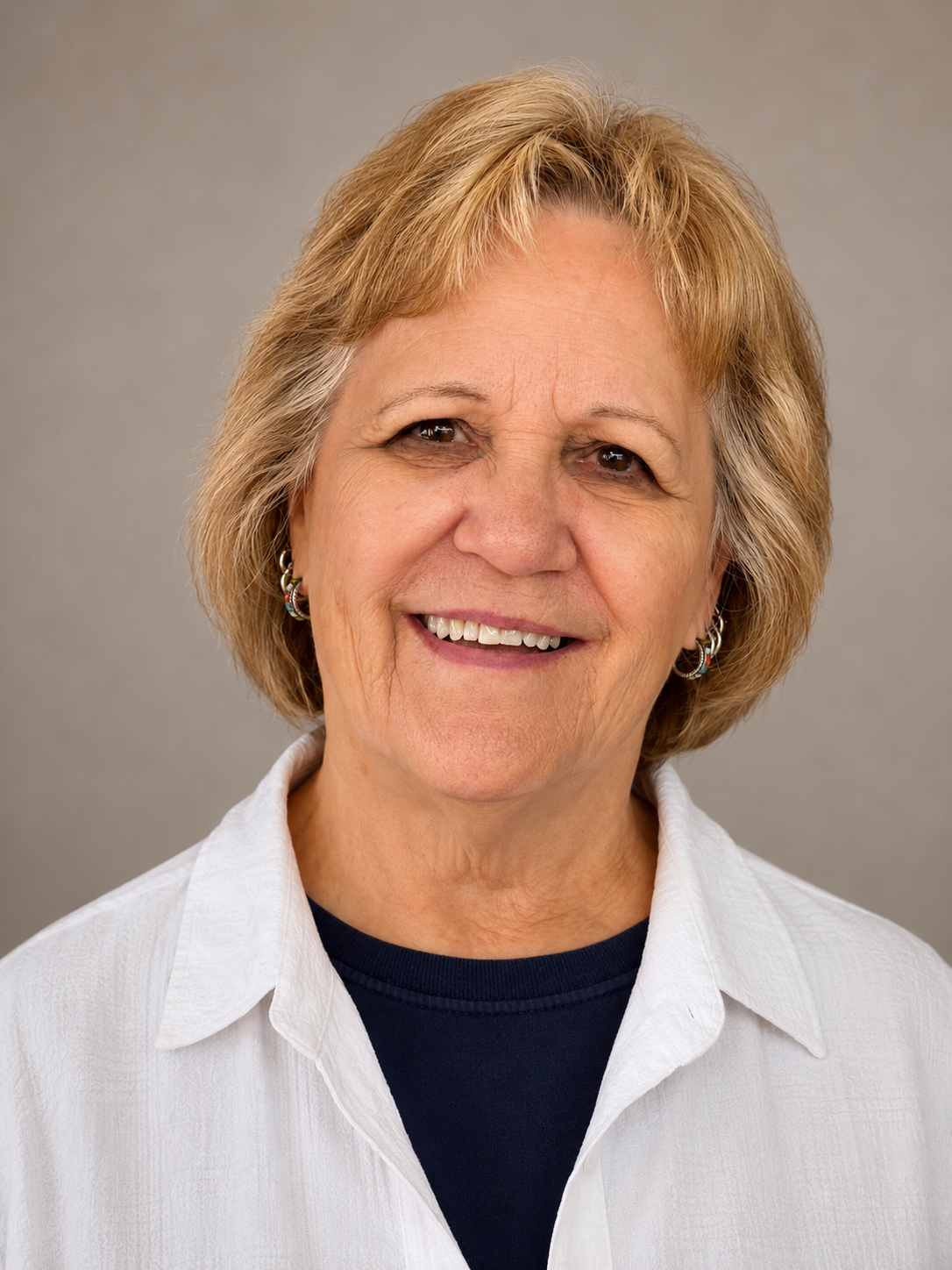 Smiling older woman with light brown hair, wearing earrings, a white shirt, and a dark top underneath, against a neutral gray background.