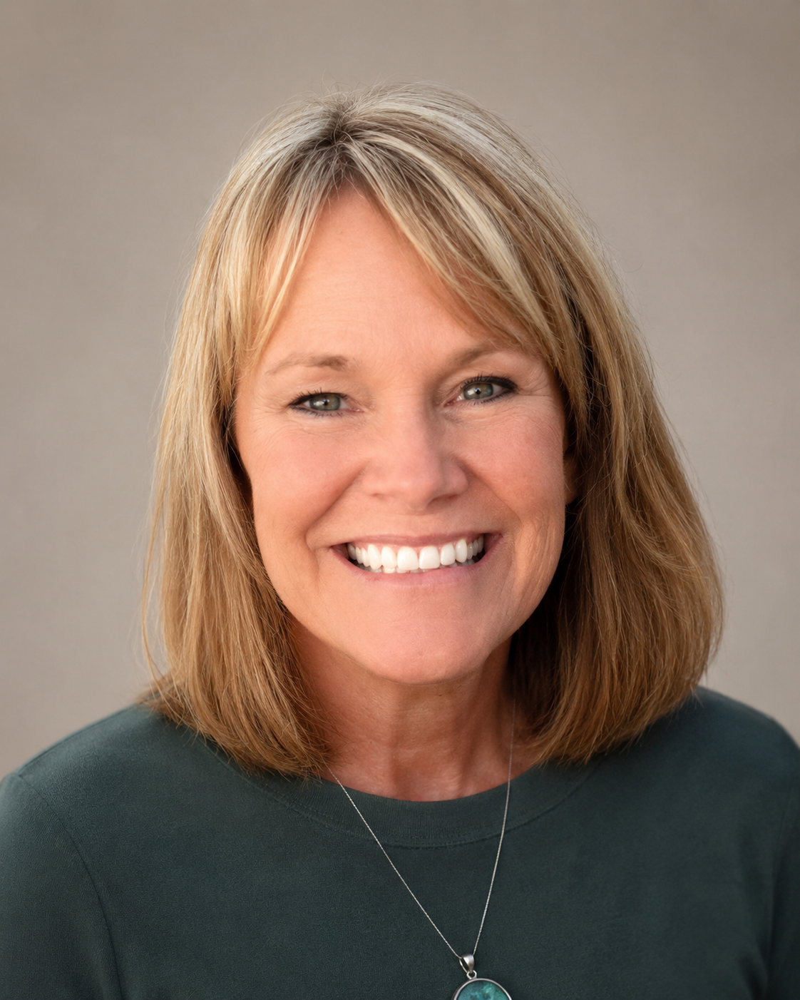 Close-up portrait of a smiling woman with shoulder-length blonde hair, green eyes, wearing a dark green top and a silver necklace with a blue pendant.