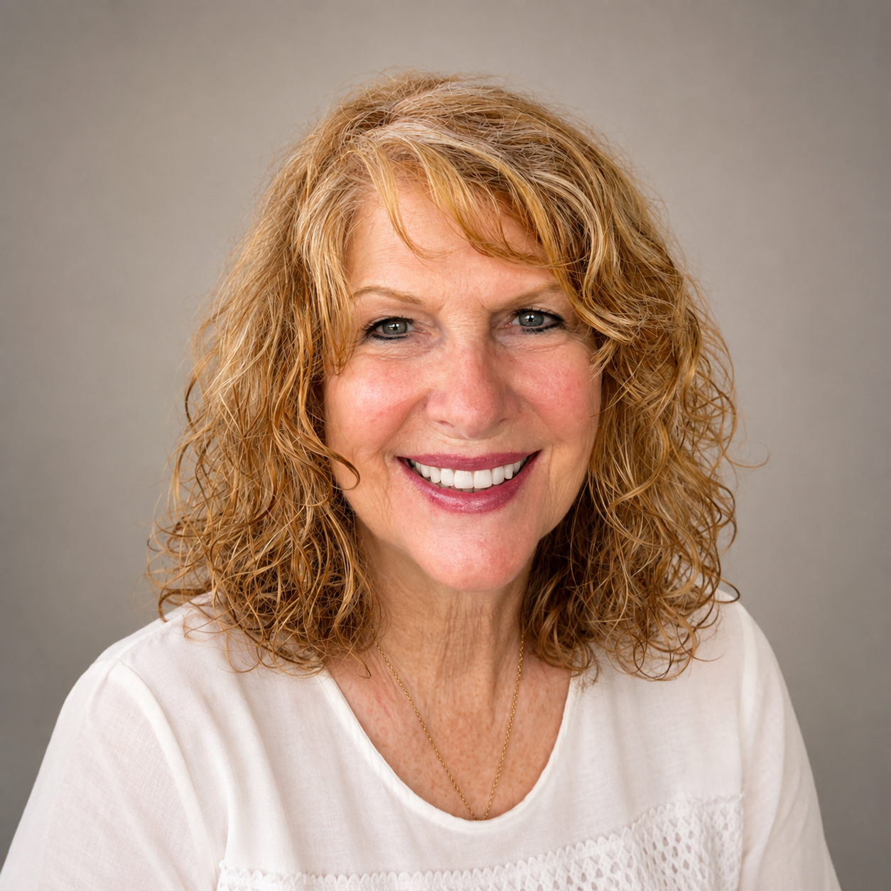 A smiling woman with curly blonde hair wearing a white top and a gold necklace against a neutral background.