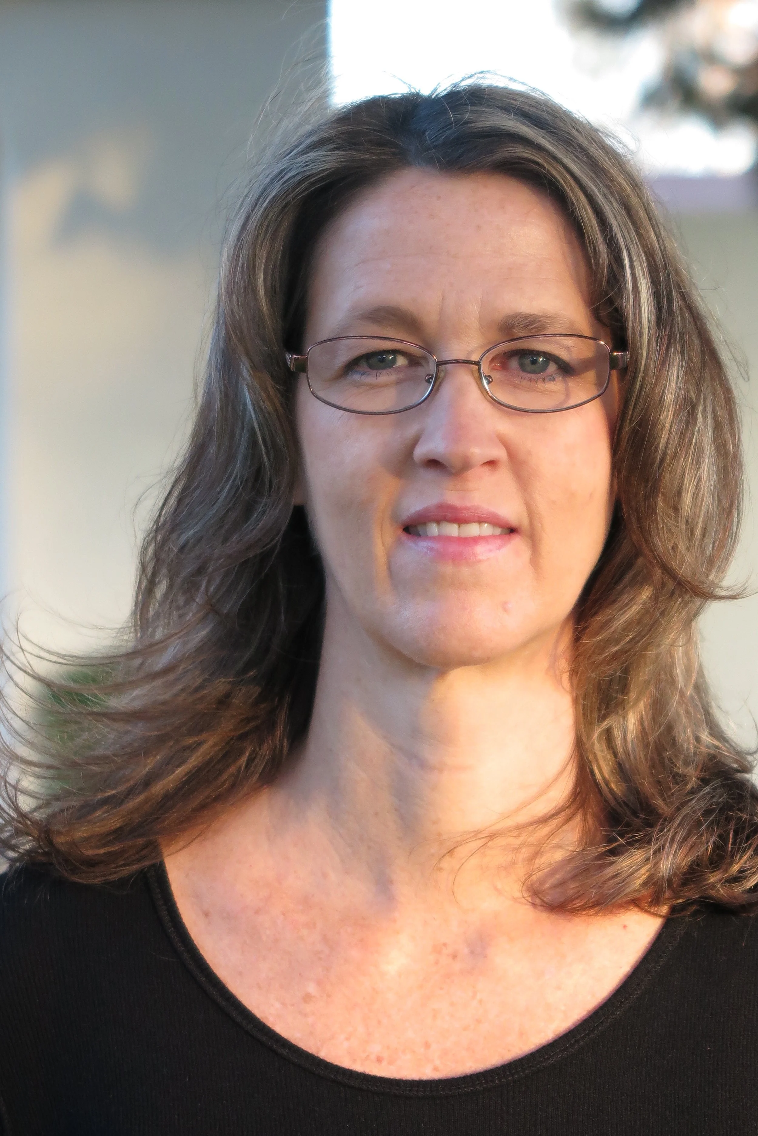 A woman with shoulder-length brown hair, wearing glasses and a black top, standing outdoors in natural light.