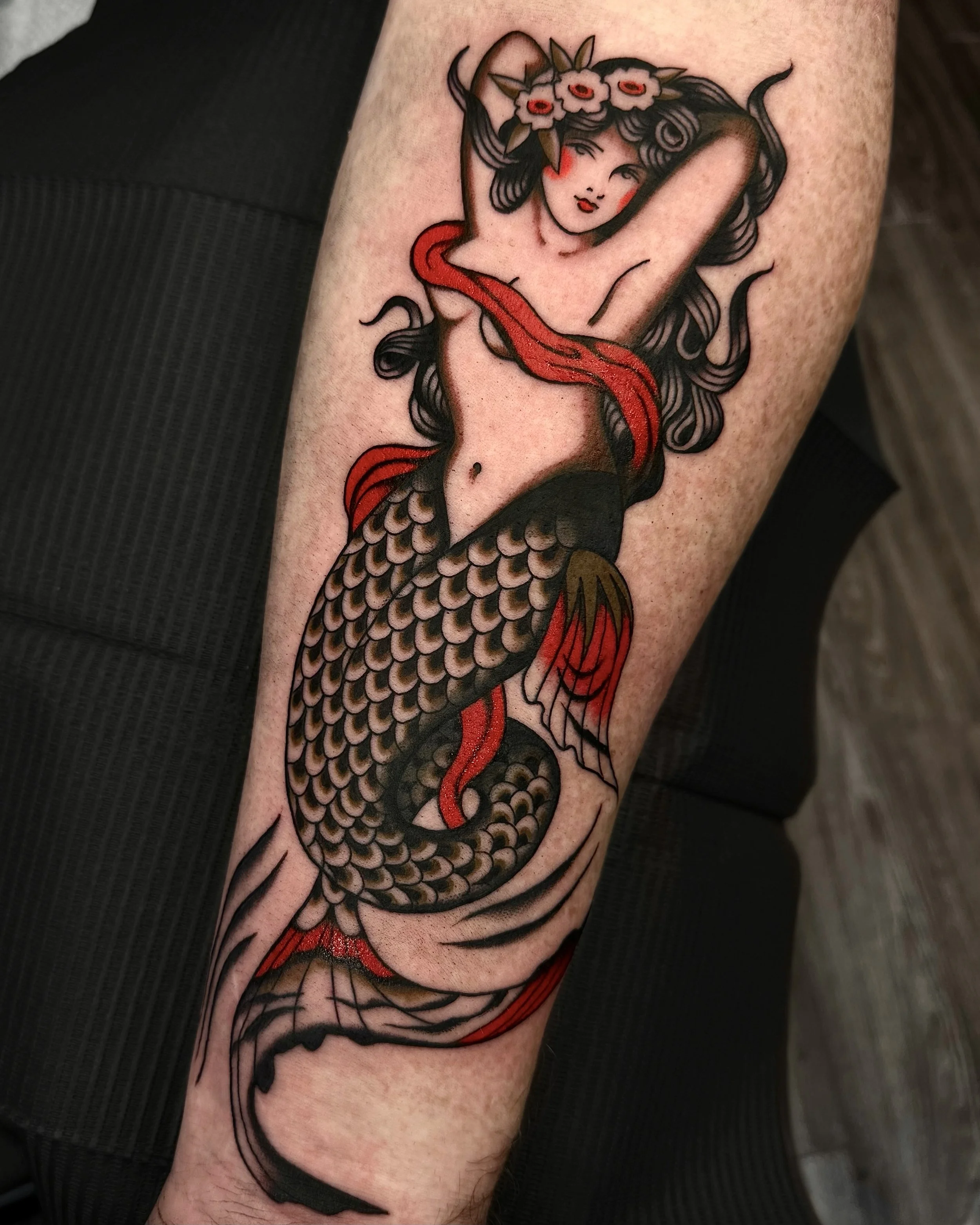 Tattoo of a mermaid with long black hair and a flower crown on a person's arm.