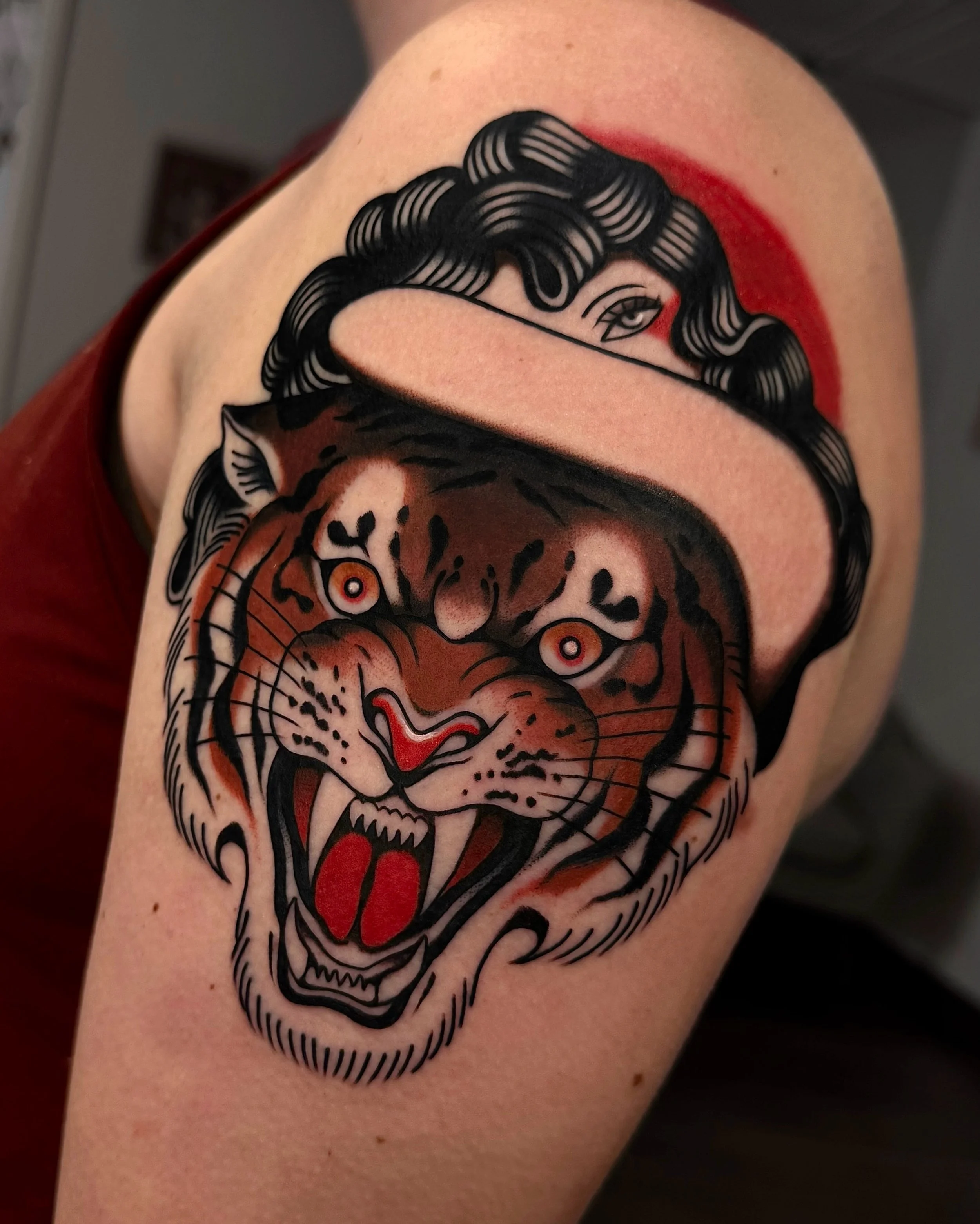 Tattoo of a roaring tiger's face with sharp teeth and red tongue, partly covered by a stylized woman’s face with curly hair and one eye visible, using traditional tattoo style with bold lines and limited colors, on person's upper arm.