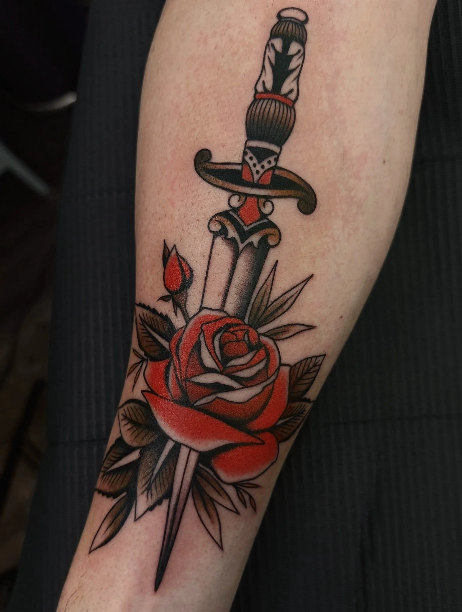 Tattoo of a sword piercing a red rose with leaves, outlined in black with red accents.