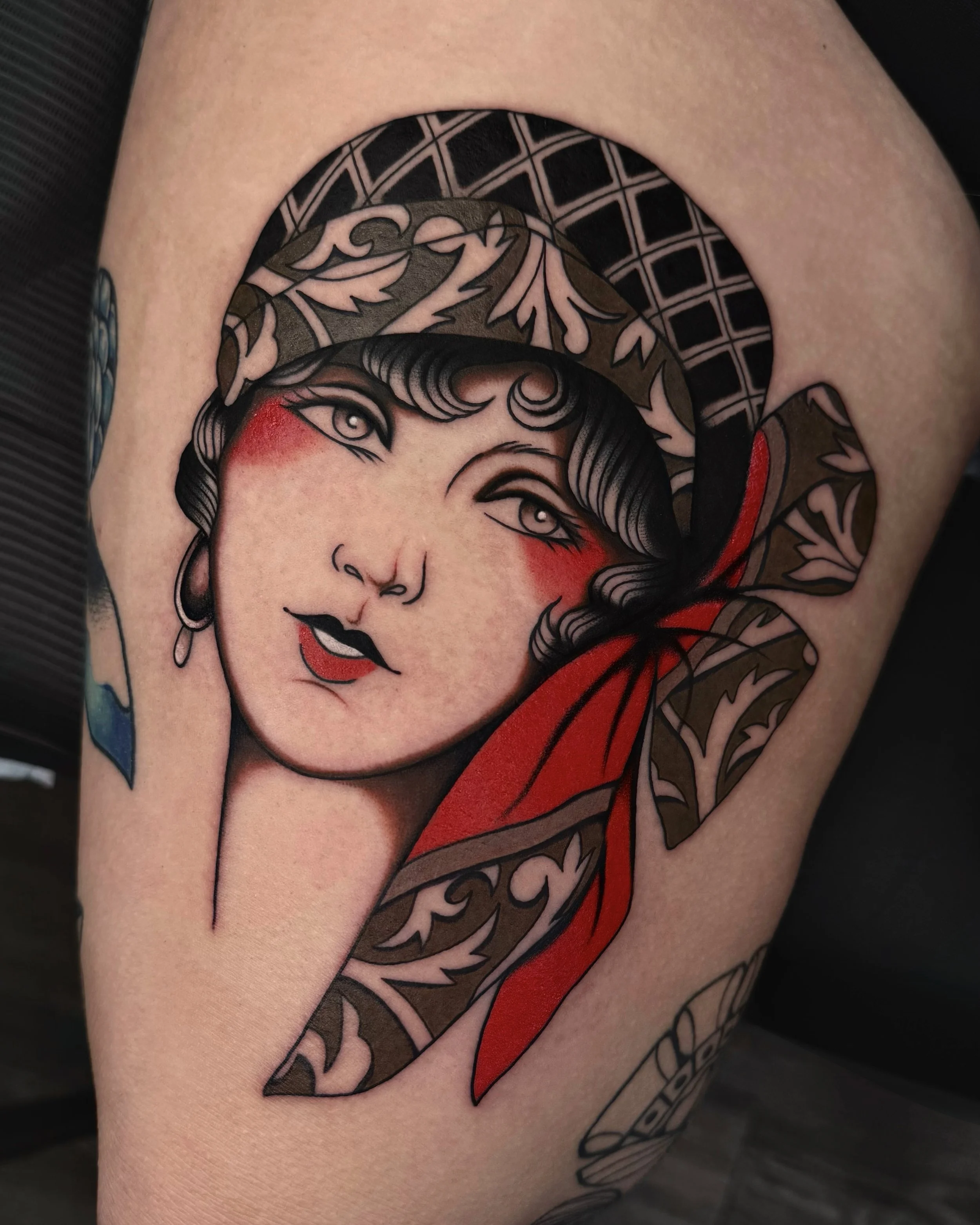 Tattoo of a woman's face with makeup, red cheeks, earrings, and styled hair, adorned with a large red and patterned brown ribbon or bow.