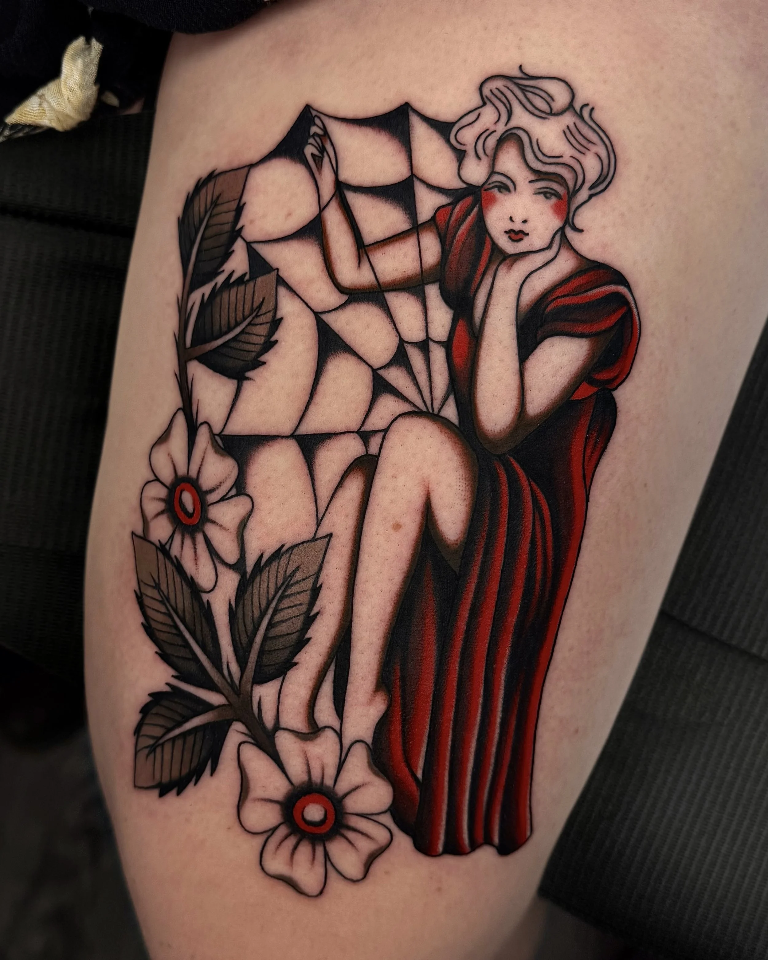 Tattoo of a woman with short, wavy hair wearing a red and black striped dress, sitting with her knees up, resting her chin on her hand, surrounded by flowers and a spider web.