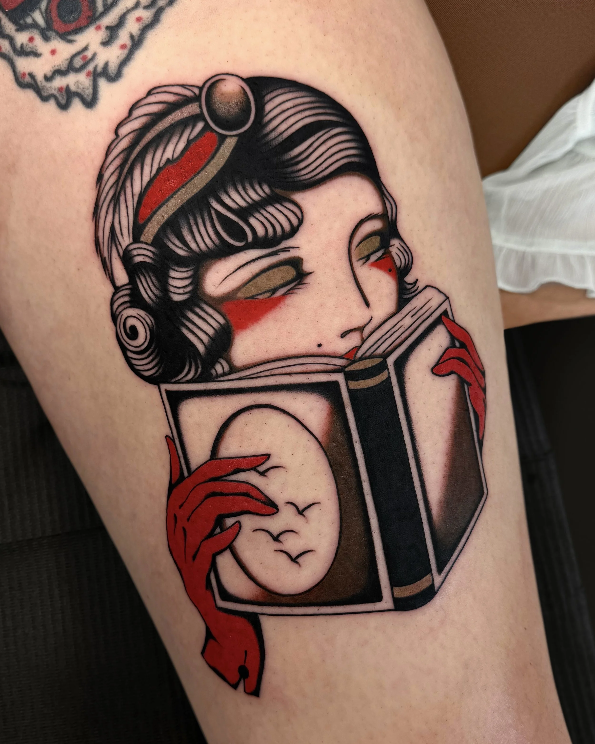 Tattoo of a woman with black hair reading a book, with red and black gloves, and stylized eye makeup, in a traditional tattoo style.