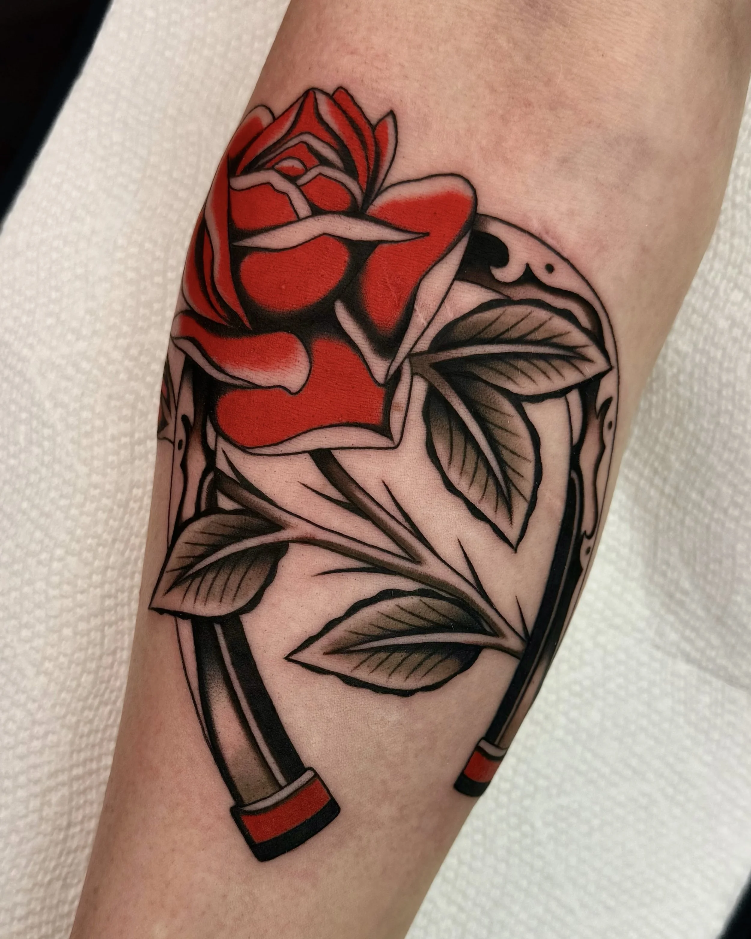 Tattoo of a red rose with green leaves and a black stem on someone's arm.