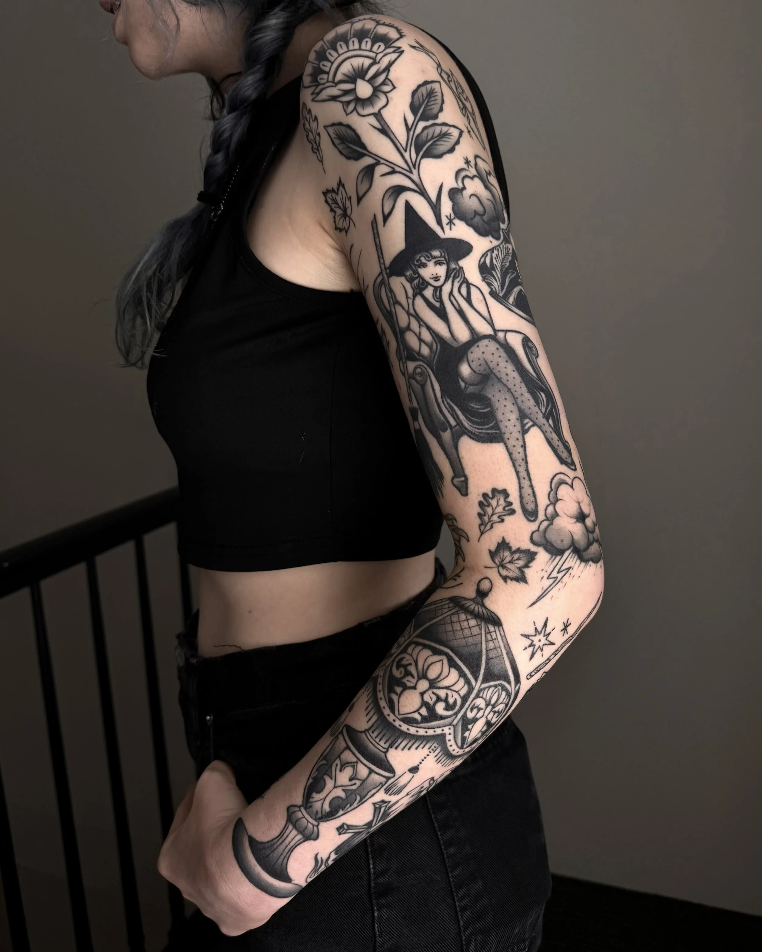 A black-and-gray tattoo sleeve of spooky imagery. The full tattoo sleeve contains a pin-up woman dressed as a witch, traditional ornamental florals, a vintage lamp, storm clouds, and fall leaves. The tattoo is done in full black
