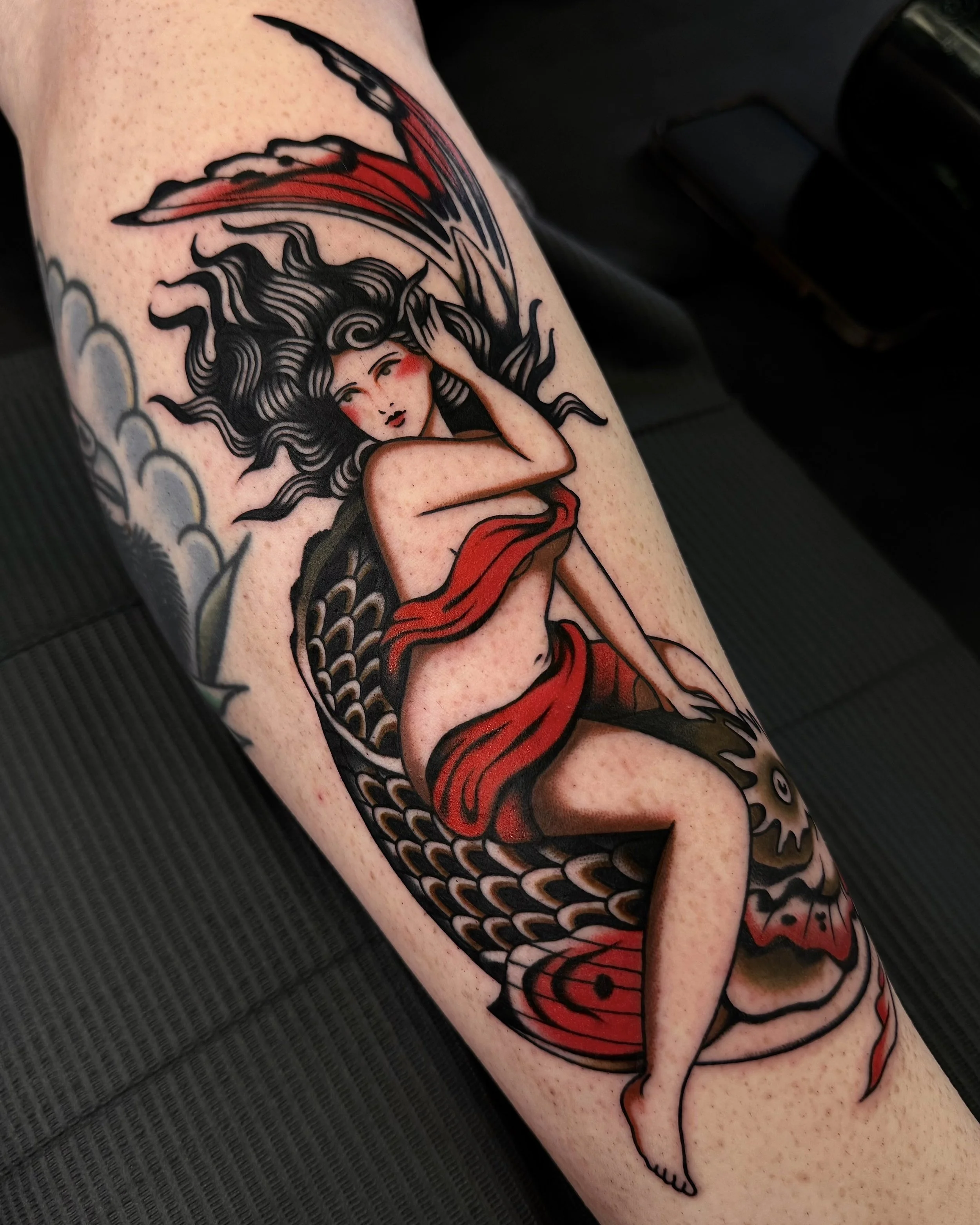Tattoo of a woman with long black hair, red markings on her face, and a red sash, sitting on a large fish with detailed scales. The woman's pose and the fish's details are stylized and traditional in appearance.