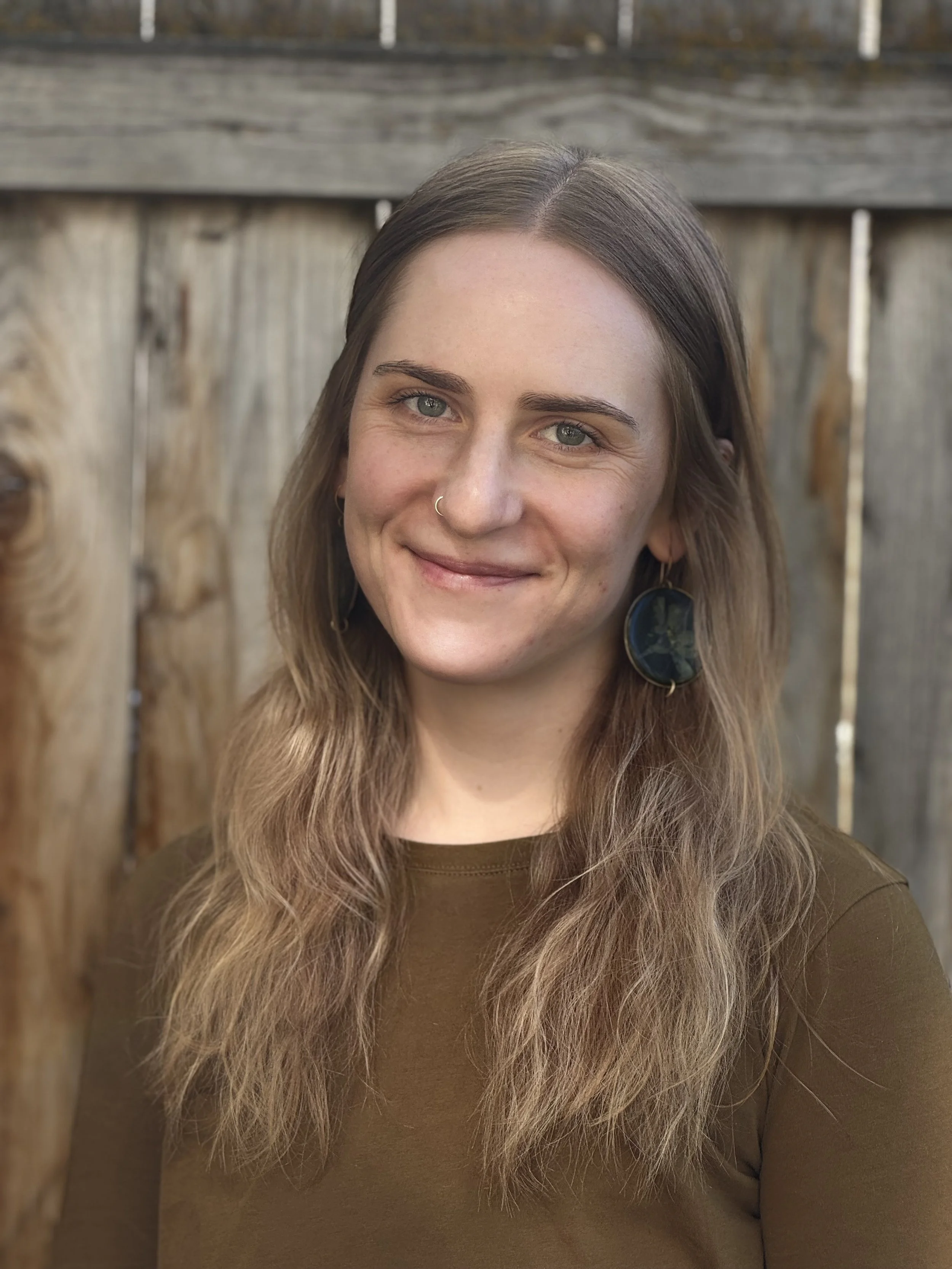 A young woman with long wavy hair, blue eyes, and a nose ring, smiling outdoors against a wooden fence, wearing a brown top and circular earrings.
