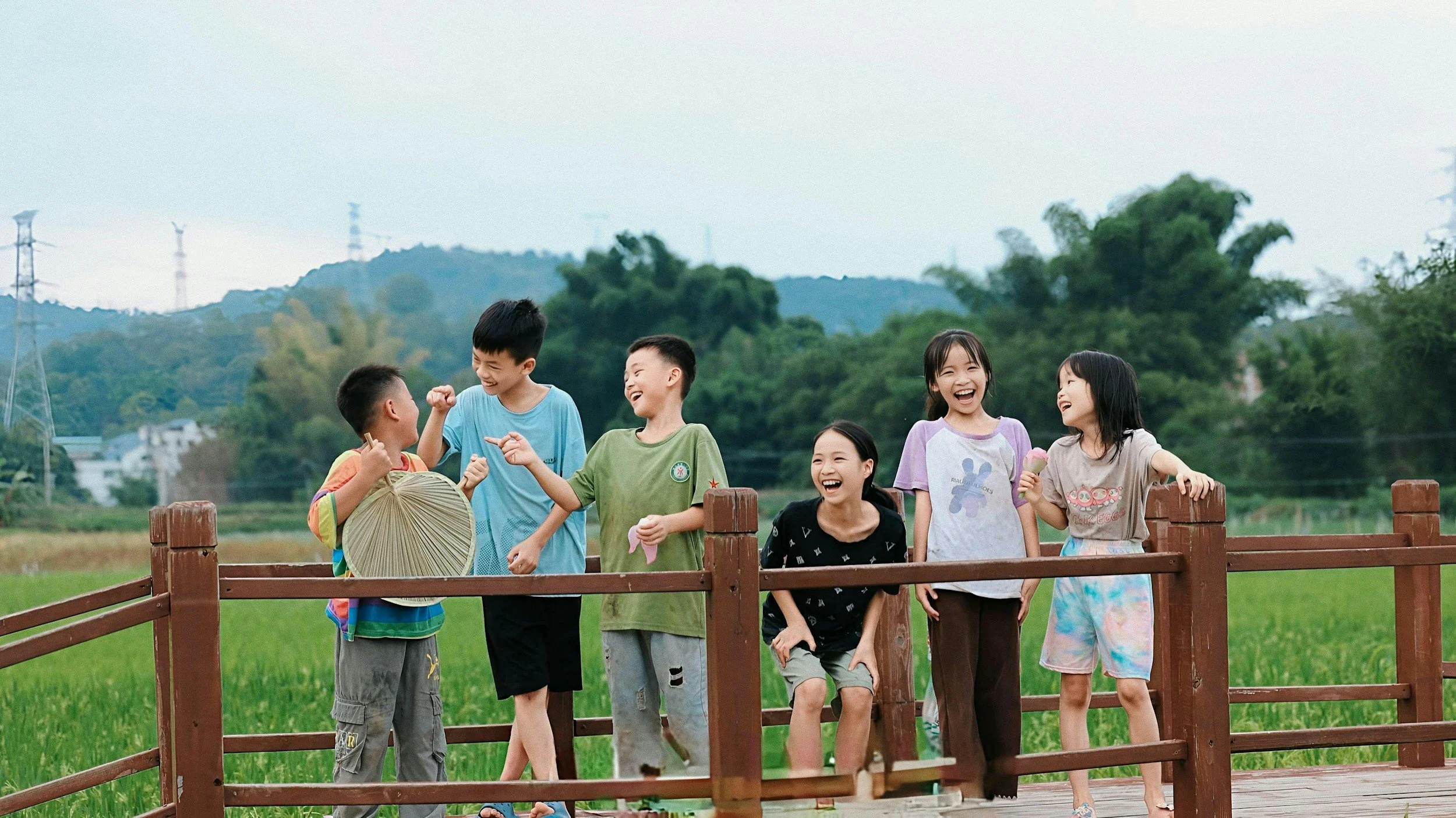 Group of children laughing and talking outdoors on a bright day, standing behind a wooden fence with green fields and trees in the background.