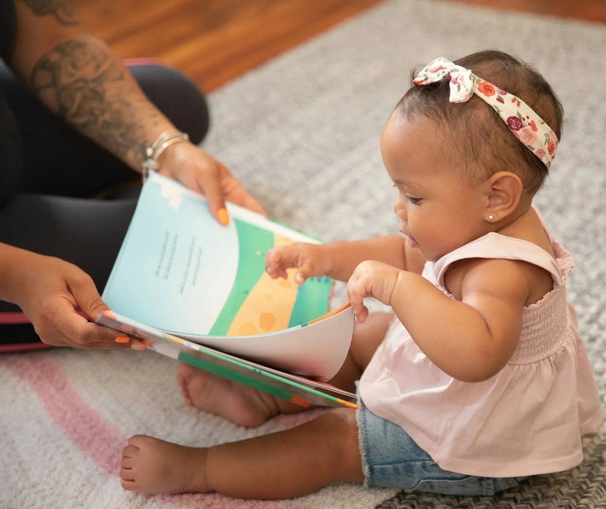 A young girl with a floral headband looks at a colorful booklet held by an adult with tattooed arms, sitting on a carpeted floor.