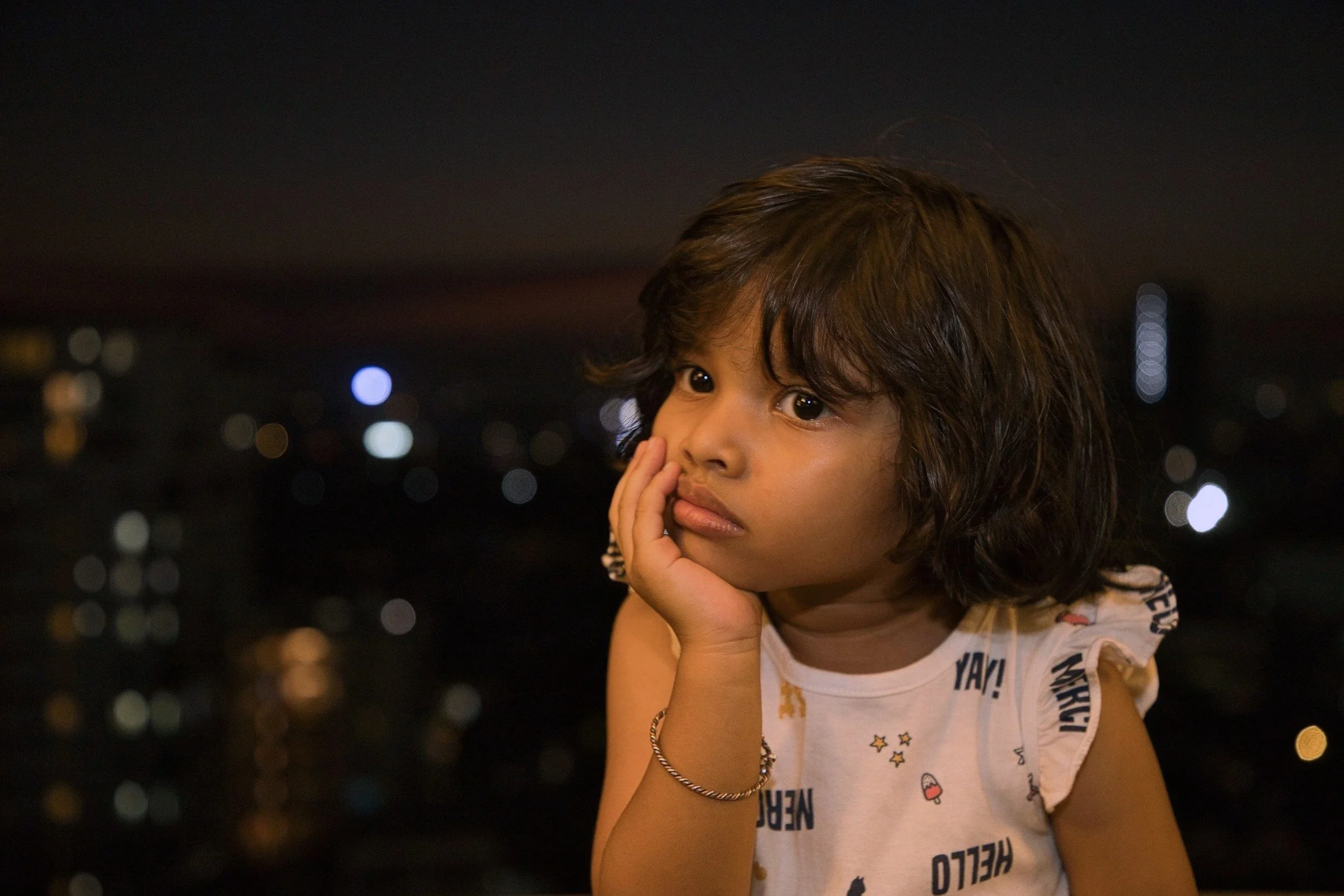 A young girl with curly brown hair and dark eyes resting her chin on her hand, looking thoughtfully at the night cityscape with blurred lights in the background.