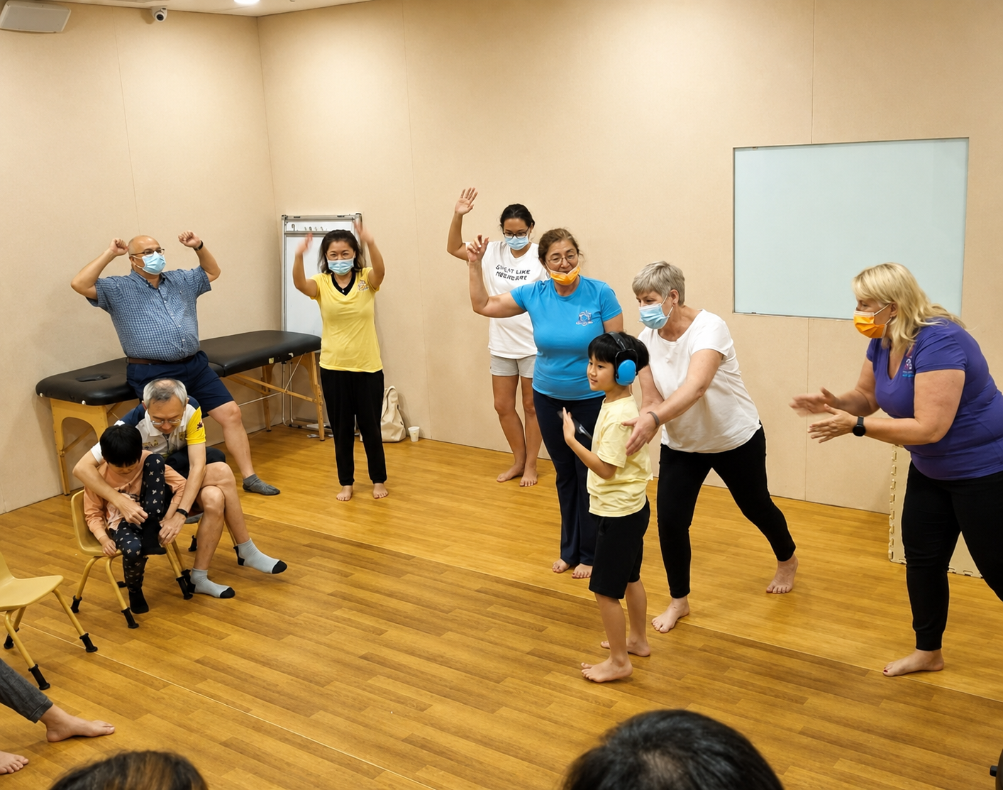 Group of people in a room, some standing and some sitting, engaging in activities and conversation. All are wearing masks, and there is a mix of children and adults.