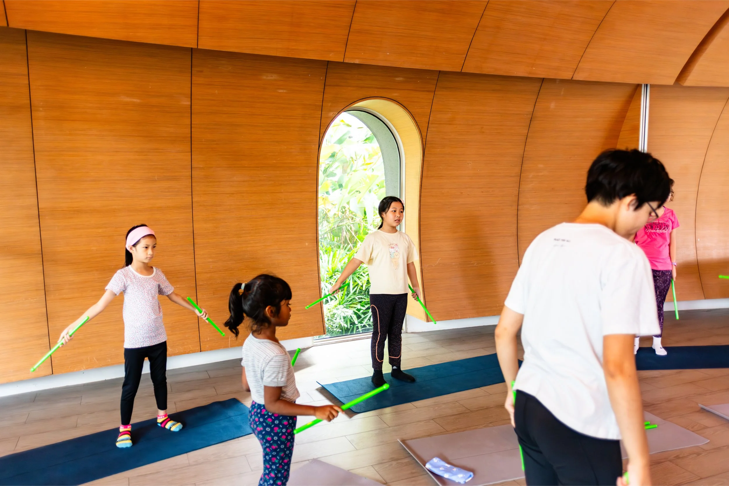 Children participating in a group activity indoors, using green sticks, with a wooden wall and a window showing greenery outside.