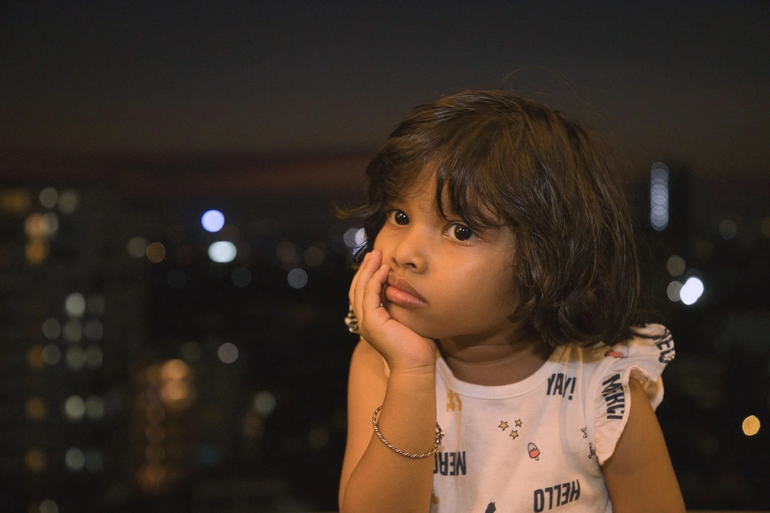 Young girl with curly brown hair, resting her chin on her hand, looking thoughtful against a blurred city skyline at night.