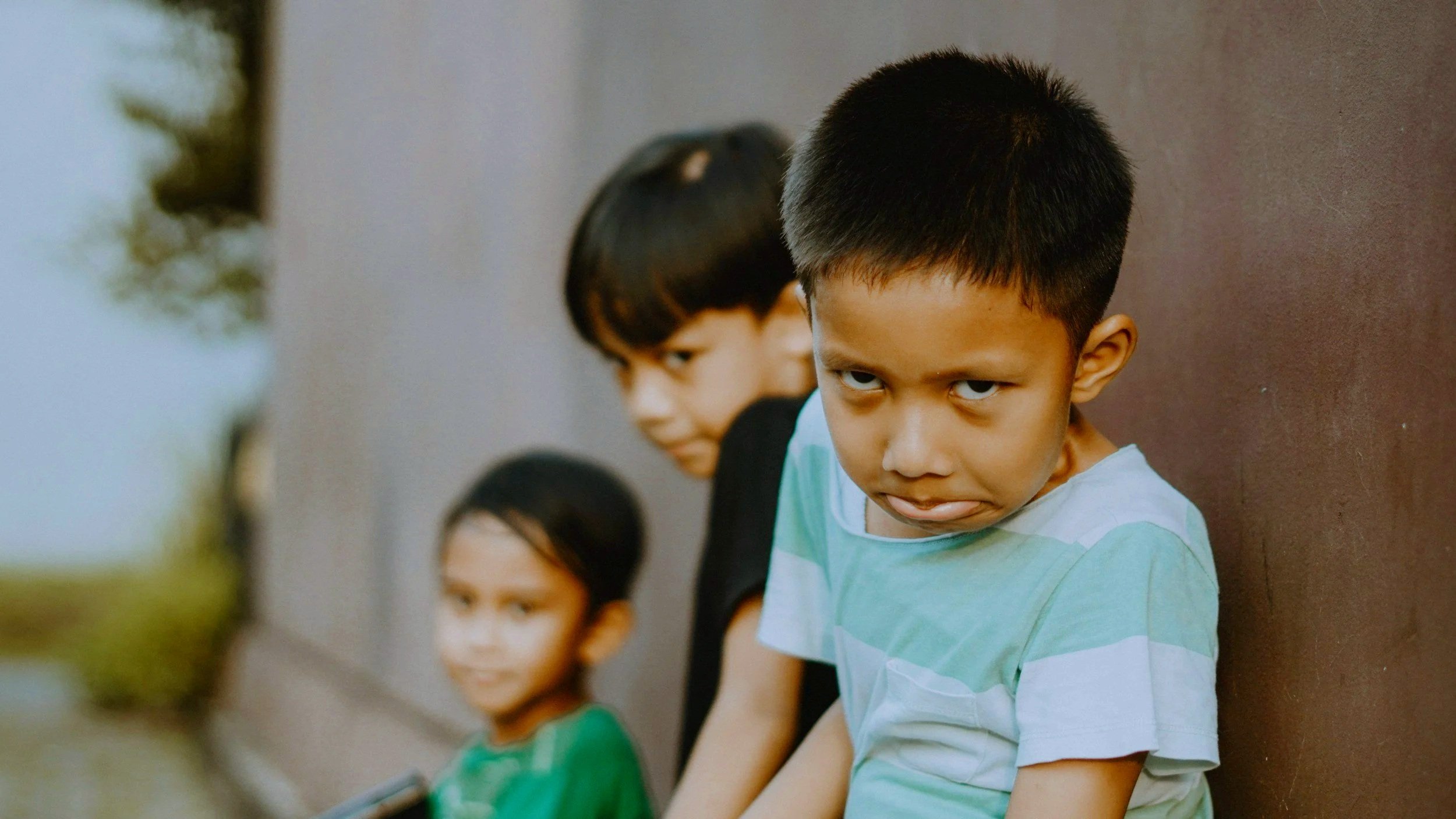 Three children standing against a wall, with one boy in the foreground looking intensely at the camera, and two girls in the background.