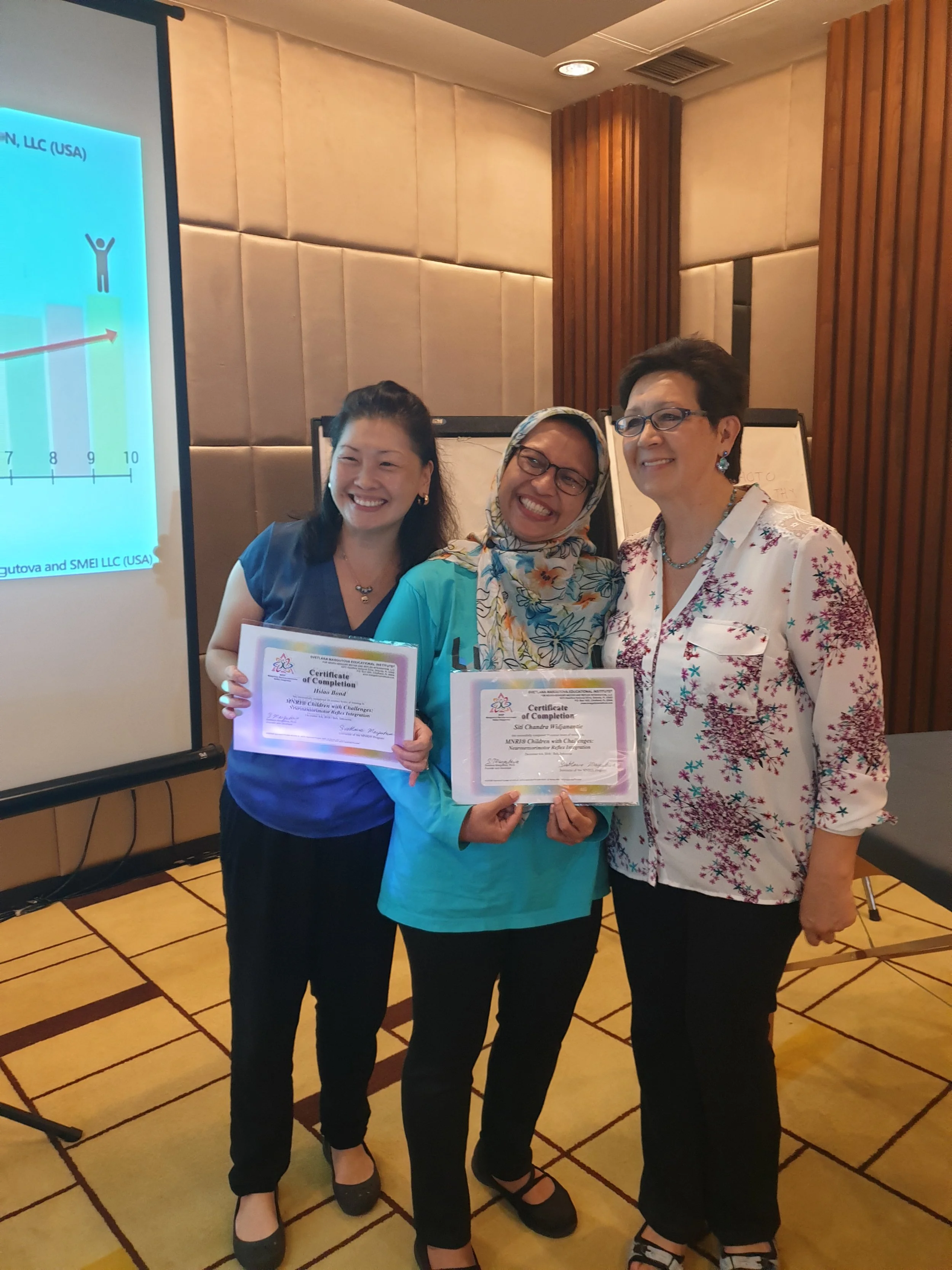 Three women standing together in a conference room, holding certificates of completion and smiling. One woman is wearing a blue top with a scarf, the second is in a white floral blouse, and the third is in a dark top. The background features a presentation screen and wooden paneling.