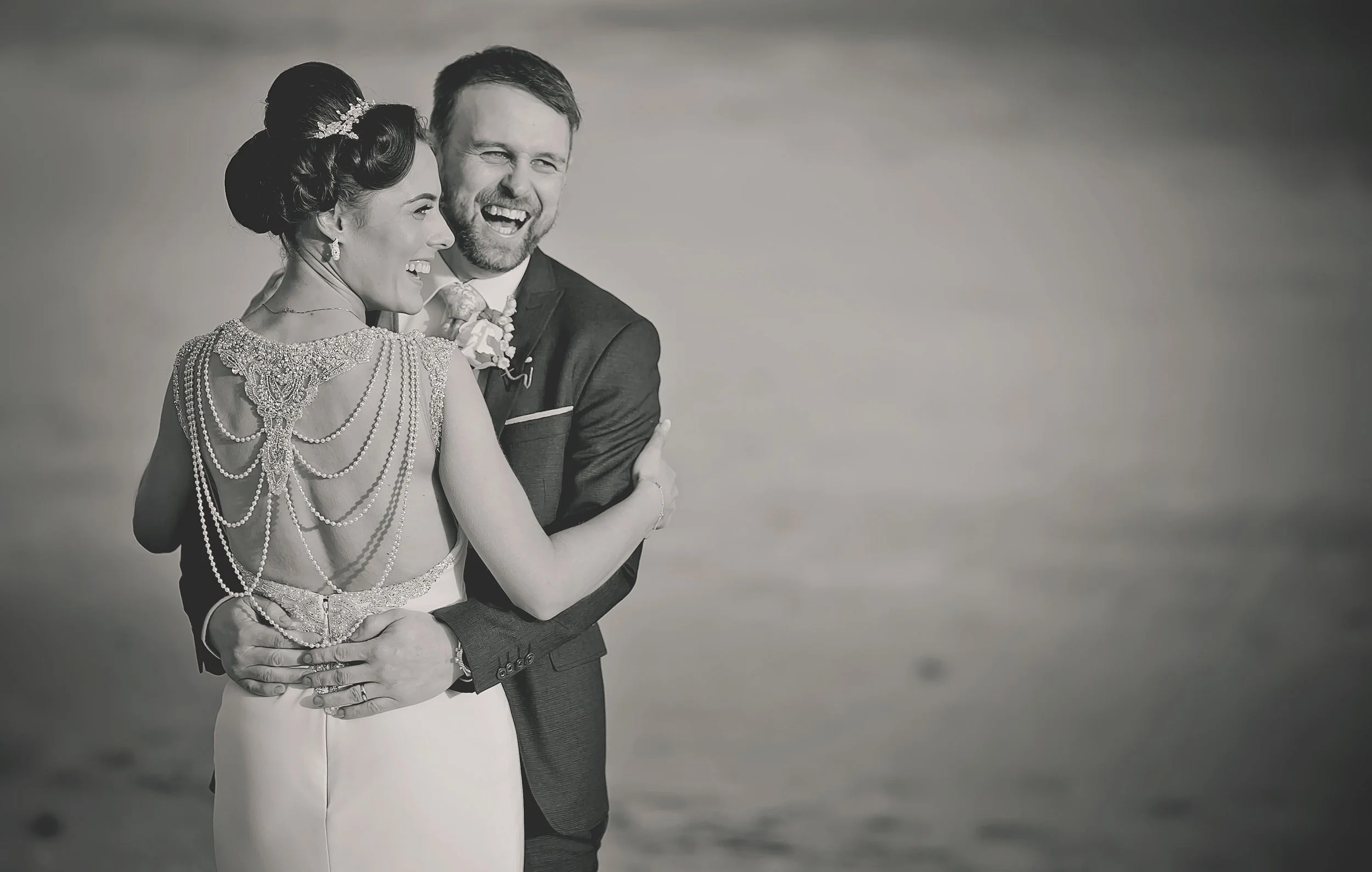 bride and groom embrace and laugh in this black-and-white portrait on the beach