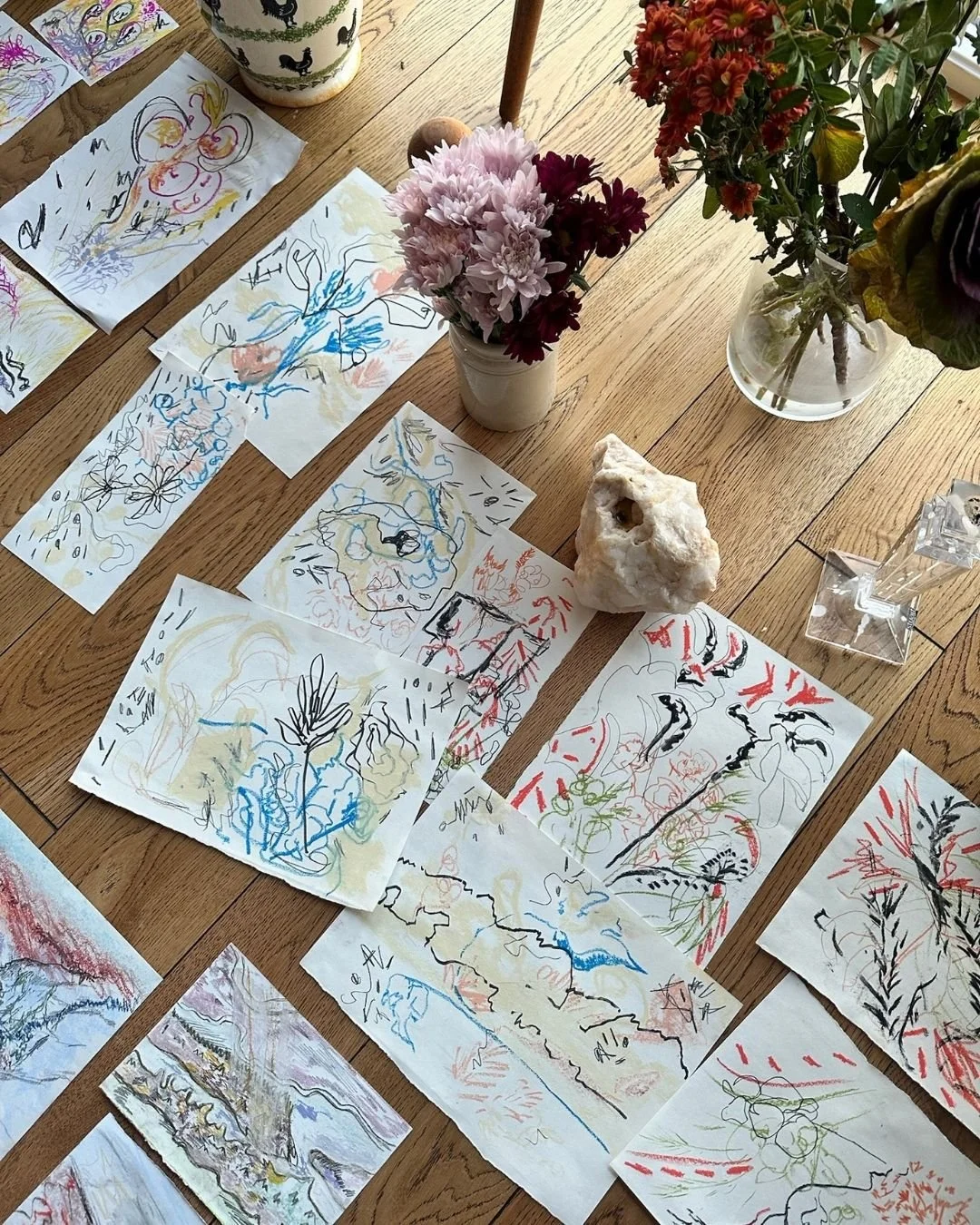 A collection of abstract, colorful sketches and paintings spread out on a wooden floor, with vases of flowers and a decorative mug nearby.