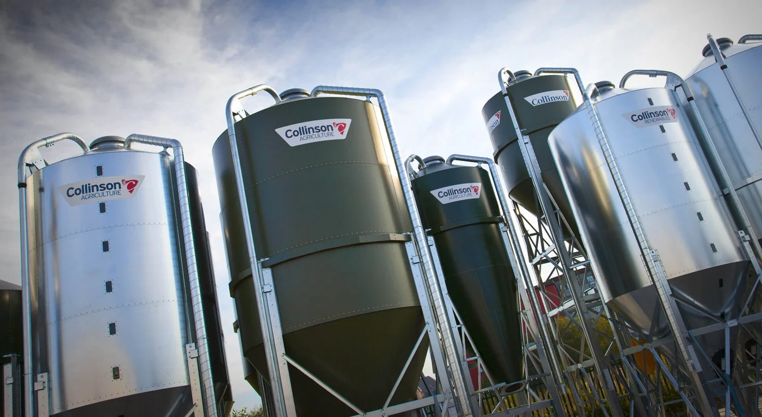 A bank of galvanised and dark green Collinson feed silos standing against a blue sky on a farm