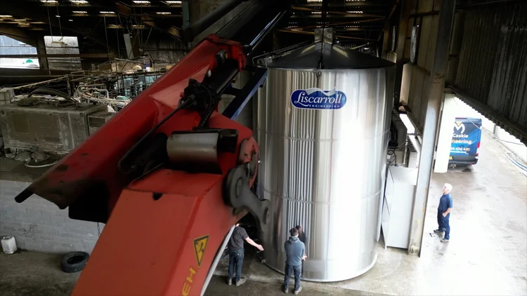 The stainless Liscarroll silo standing upright under a covered yard, a lorry-mounted crane folded beside it and two people at its base