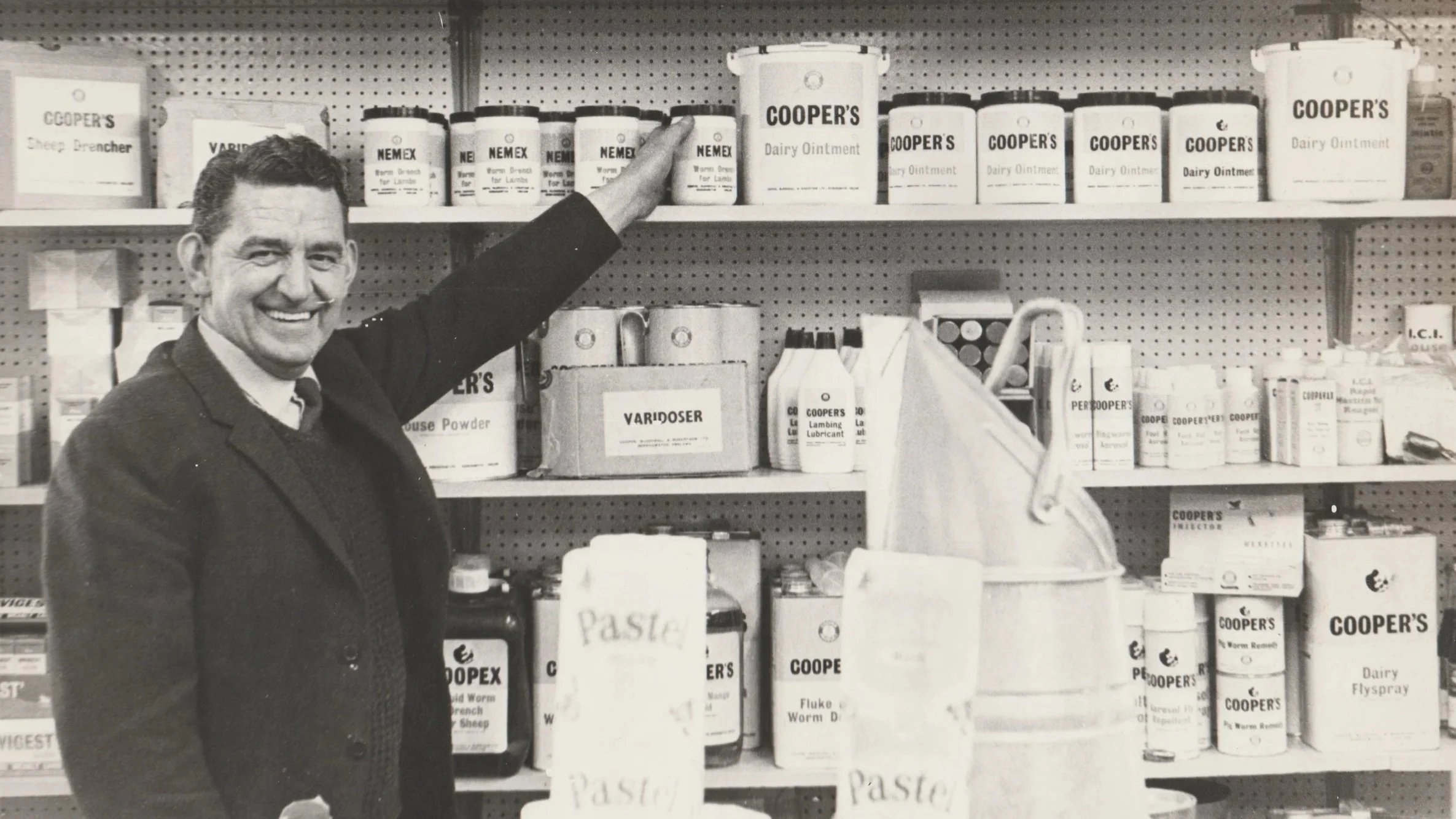 A man in a suit and tie smiling and pointing at shelves filled with vintage pharmacy or hardware products, including ointments, powders, and cans labeled with brand names like Cooper's and Nemex, in a black-and-white photo.
