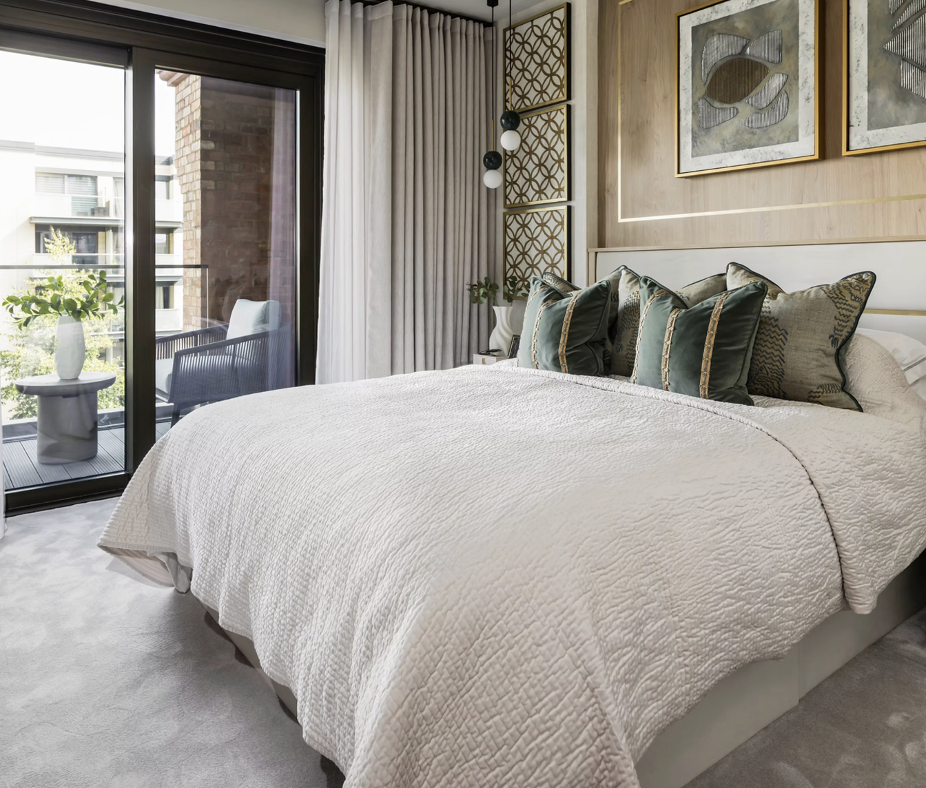 A modern bedroom with a large bed covered in a white textured quilt, decorated with multiple dark green and patterned pillows. There is a small balcony with outdoor seating and a potted plant visible through sliding glass doors. The room features light-colored curtains, art on the wall, and a neutral-color carpet.