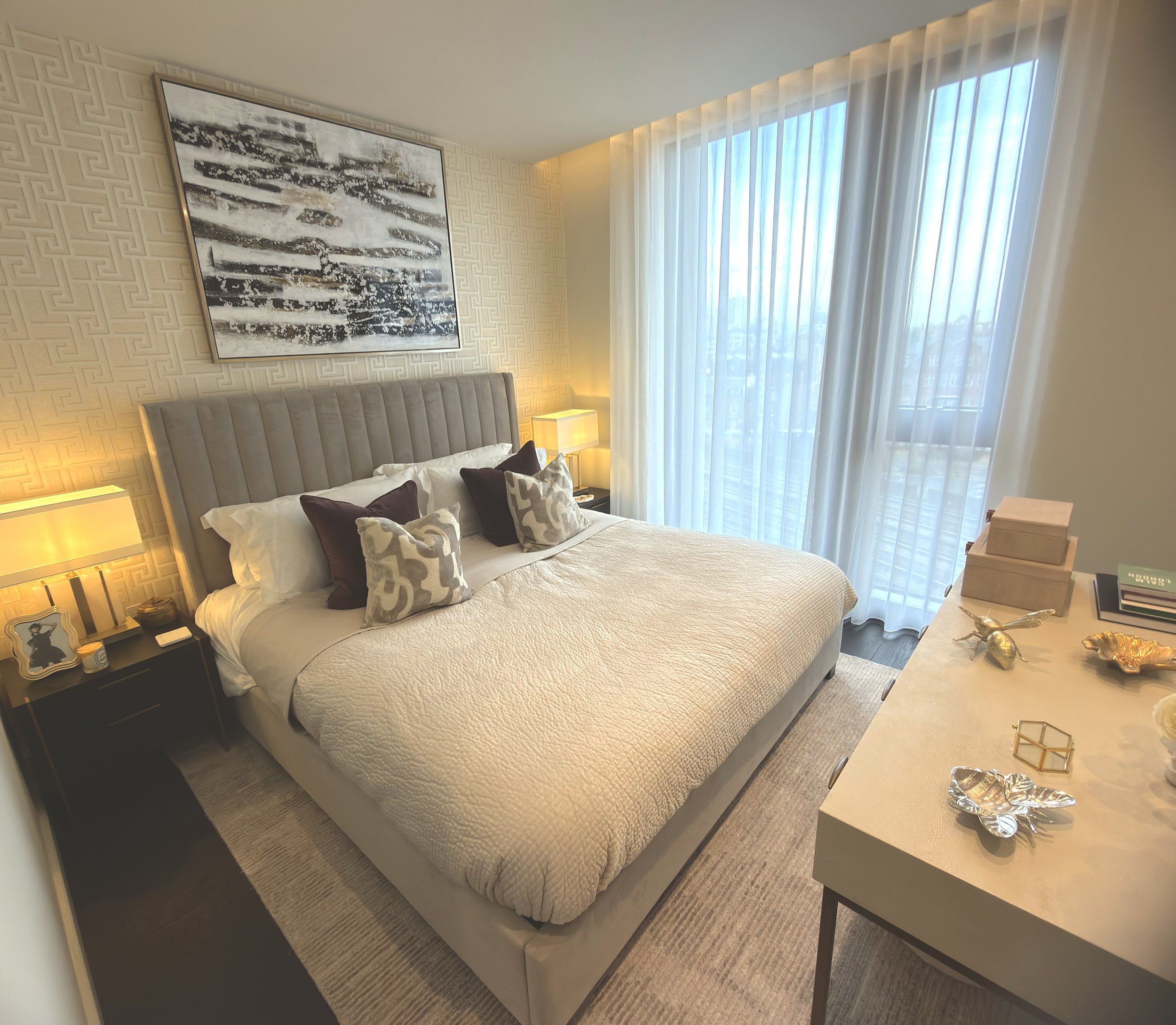 A modern bedroom with a king-sized bed, beige quilted bedspread, and an array of decorative pillows. There are two black nightstands with beige lamps on each side of the bed, a large abstract painting above the headboard, and a window with sheer curtains letting in natural light. A dresser with decorative items and small boxes is on the right side of the room.