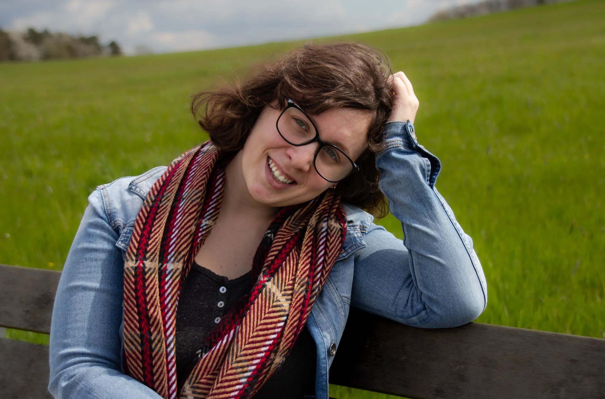 Junge Frau mit Brille, dunklem Haar, lächelnd, auf einer Parkbank, im Grünen, trägt Jeansjacke, bunte Schal, im Freien.