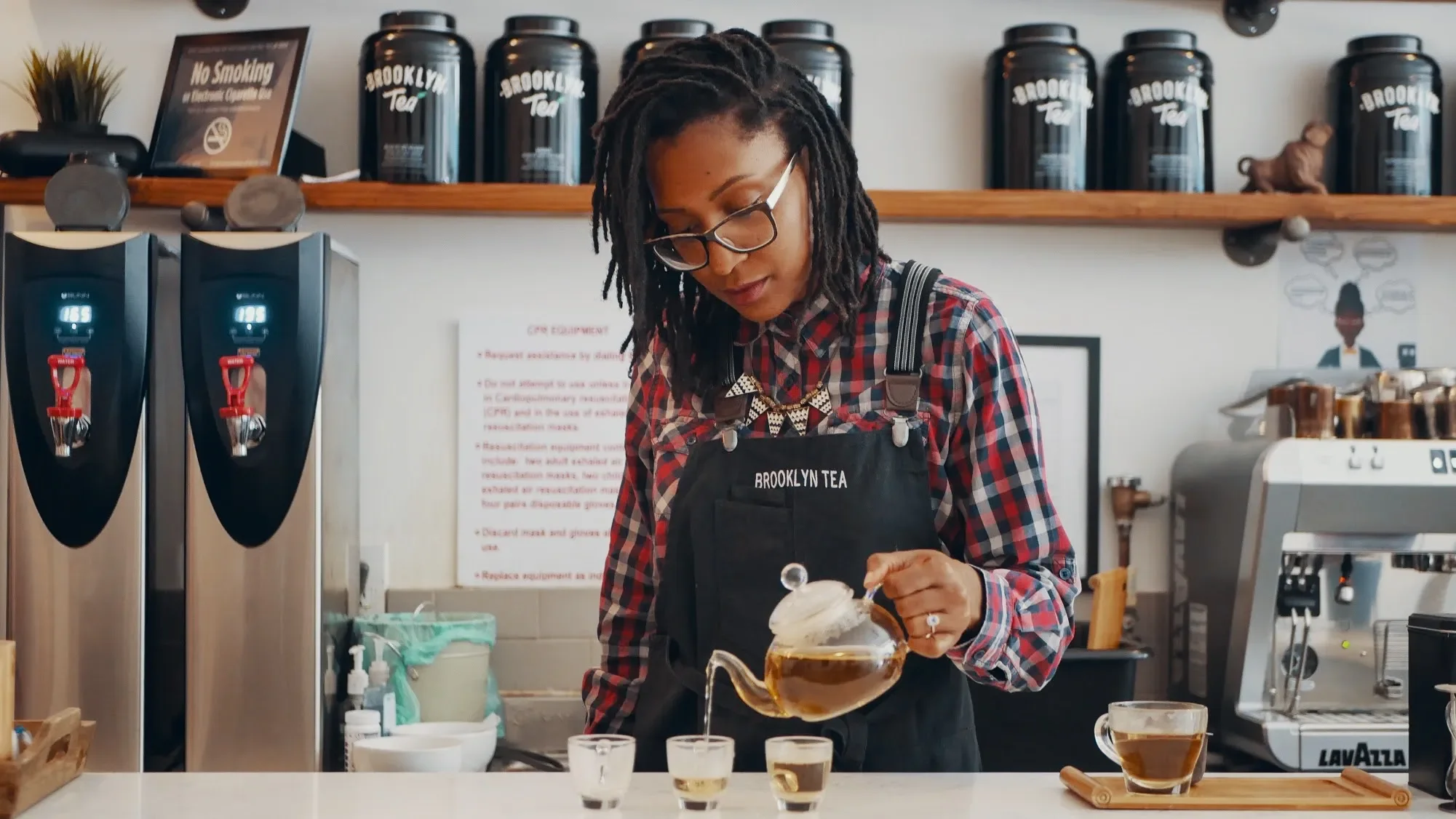 A woman with dreadlocks, glasses, and a Brooklyn Tea apron pours tea from a teapot into small glass cups in a tea shop.