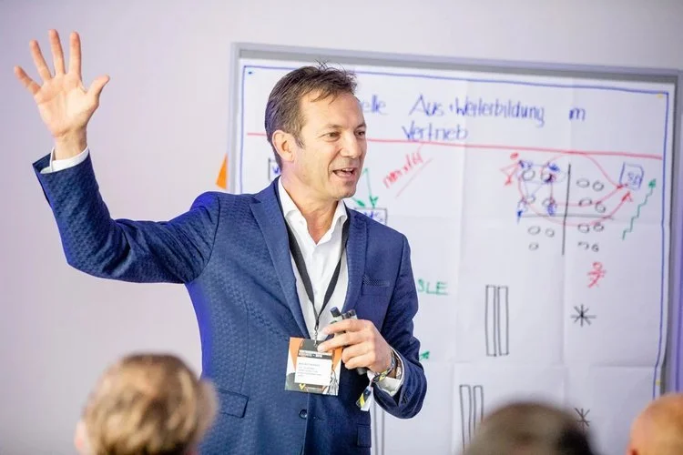 Mann in blauer Jacke hält Mikrofon und erklärt eine Whiteboard-Skizze.