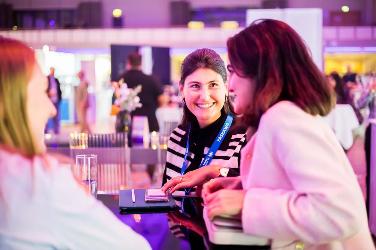 Drei Frauen unterhalten sich bei einem gesellschaftlichen Ereignis in einem hell erleuchteten Raum mit Dekorationen und Blumen, während sie an einem Tisch sitzen.