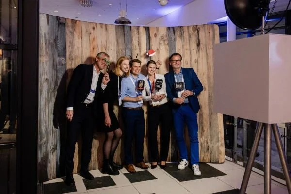 Fünf Menschen posieren auf einer Fotowand bei einer Feier. Sie tragen Name Schilder und sind in formeller Kleidung.